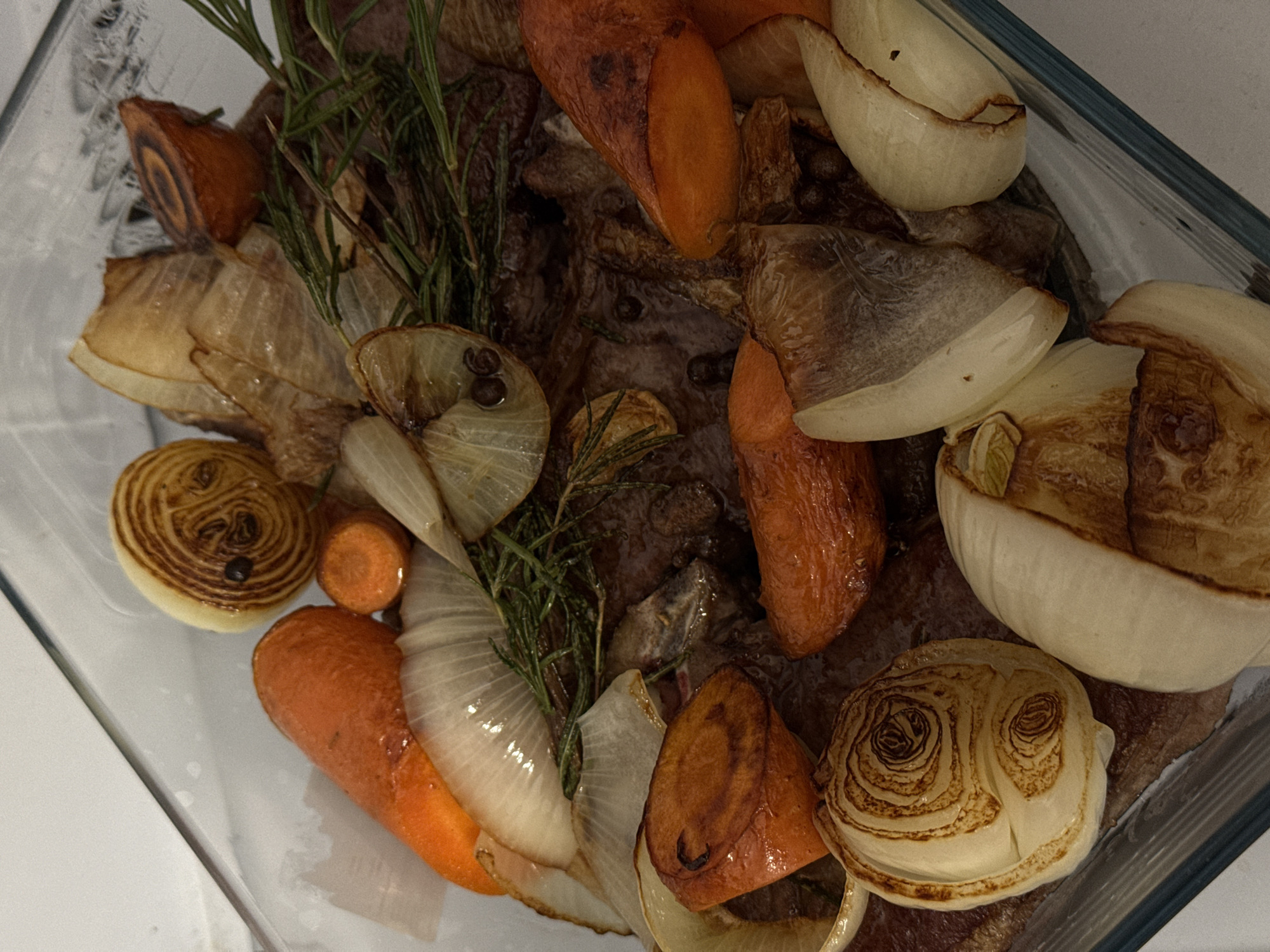The image shows a clear glass baking dish filled with roasted food. The dish contains roasted carrots, onion wedges, and what appears to be a piece of meat. There are also sprigs of rosemary visible. There is no information about the location or people in the image.
