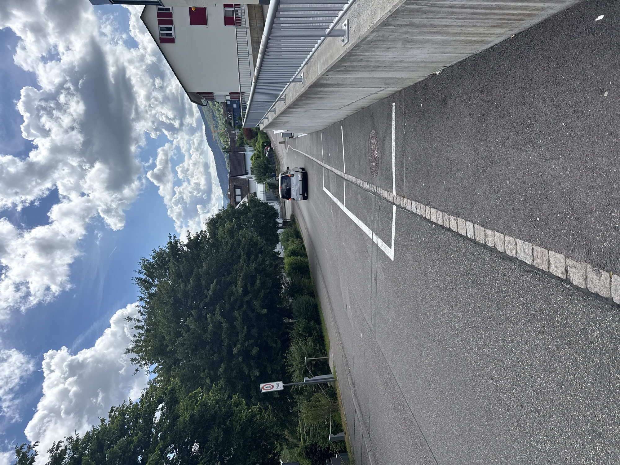 The image shows a street in Lausen, Switzerland, viewed from an elevated perspective. The street is asphalt with white painted parking spaces and a border of cobblestones along one side. A white minivan is driving away from the viewer on the street. A building with red shutters runs along the left of the road, with a concrete retaining wall and metal railing separating it from the street. A large tree borders the other side of the street. A road sign labeled "Privat" is visible along the street. In the background are distant buildings and a mountain range. The sky is blue with large cumulus clouds.
