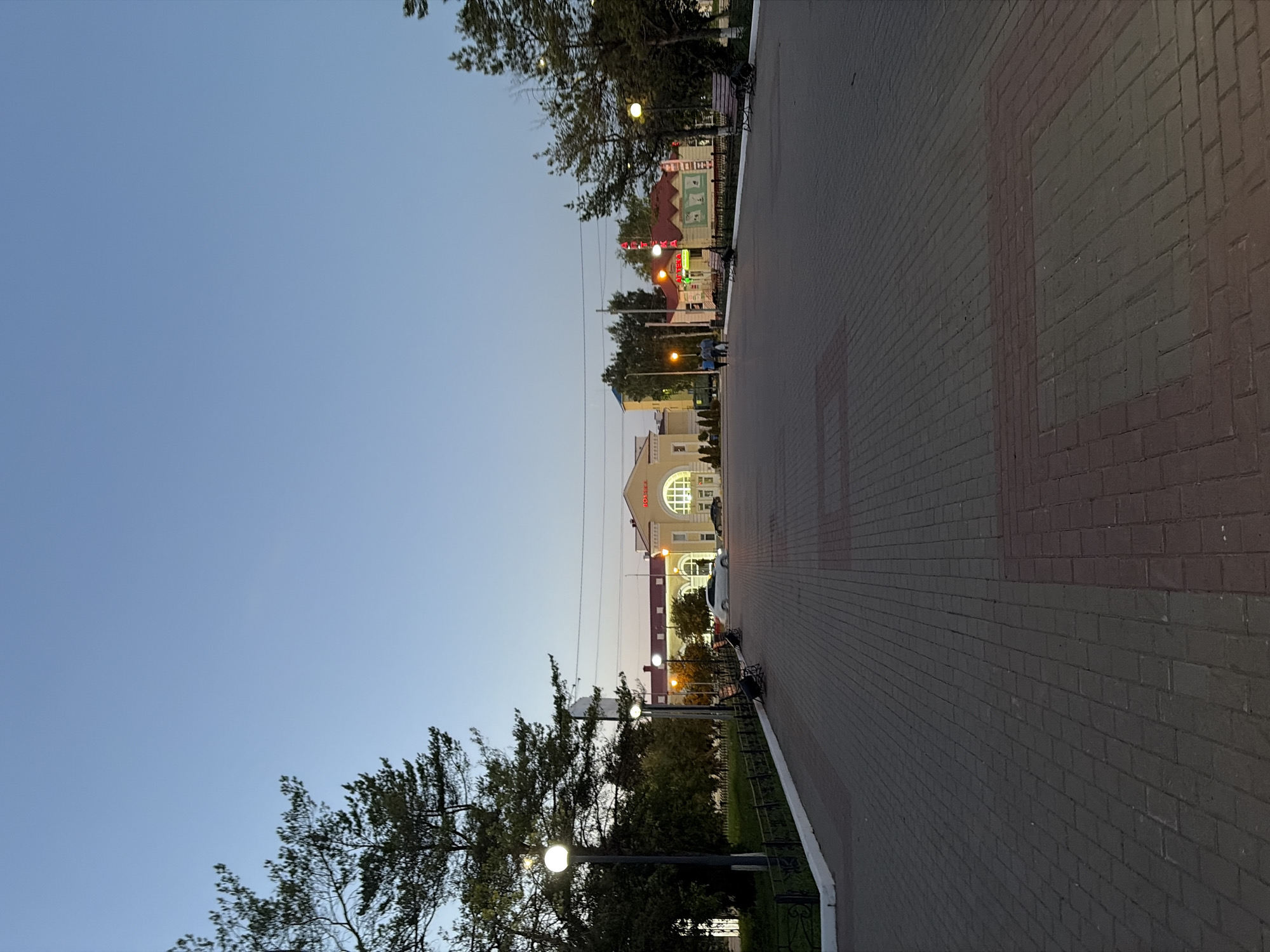 This is a medium-shot, vertical, outdoor, daylight photograph of a paved plaza in front of buildings that appear to be part of a train station or civic center in Tuymazy, Russia. The sky is clear and a pale blue, with the light suggesting early evening or late afternoon. In the foreground, a wide pathway made of brick pavers stretches towards the buildings. The pavers are laid in a pattern with rectangular insets of a different color. To the left of the pathway, there is a black metal fence with a white top edge, separating the plaza from a grassy area and trees. Several lampposts with round globes are visible along this fence.
In the middle ground, several people are walking away from the camera, heading towards the buildings. There are also a few parked cars, including a white sedan. The buildings in the background have a light-colored facade and red roofs. One building prominently features arched windows and signage that includes the word "ТУРНИКЕТ" in red illuminated letters. To the right, another building with a red roof has yellow walls and colorful signage, including the word "АПТЕКА" in red illuminated letters, indicating a pharmacy. Benches are visible along the pathway in front of both buildings. Trees are scattered throughout the scene, with their branches reaching into the frame. The overall impression is that of a public space at dusk, with some activity but not crowded.