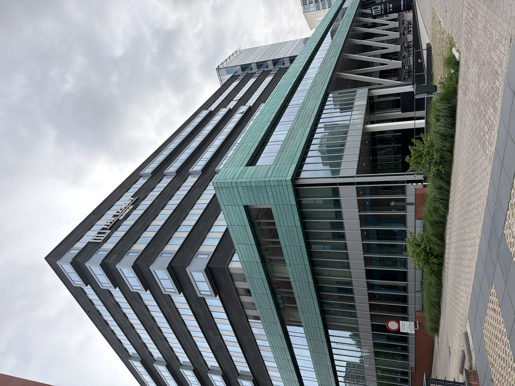 The image shows a modern building in Rijswijk, Netherlands. The building is multi-story with a dark facade on the upper levels and a green-tiled section on the middle levels. The lower levels feature extensive glass windows. The building has a prominent sign that reads "THE LOBBY" with smaller text below that appears to be the address "LANGE KLEIWEG 6-28". In front of the building, there is a paved walkway and an area with tables and chairs under a covered area. There is also some landscaping with grass and bushes. The sky is overcast.