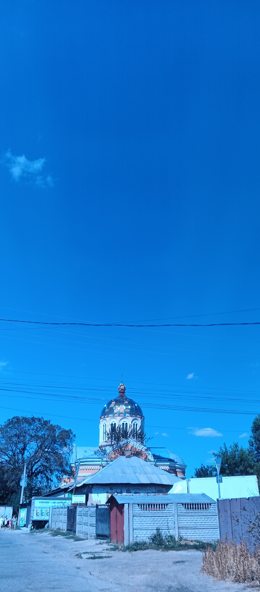 A bright blue sky dominates the upper portion of the image, with a single white cloud visible in the distance. A few power lines crisscross the sky horizontally in the middle ground. In the lower half of the image, a domed church with ornate details is visible, topped with a gold finial. In front of the church, there are several low-lying structures with corrugated metal roofs. A fence made of precast concrete panels runs across the foreground, with a small red-doored shed set against it. To the left, a large tree with dense foliage stands beside a white truck with text that appears to read "CEMARK - ЦЕ ЦЕМЕНТ". Several people are visible walking near the truck, suggesting a street or commercial area. The overall scene appears to be an outdoor daytime view in the city of Smila, Ukraine.
