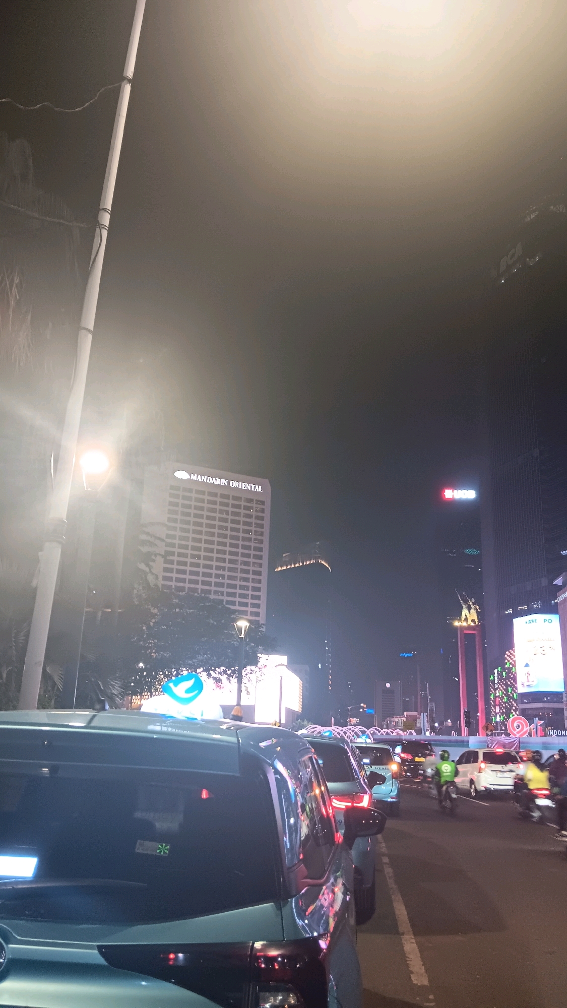 The image captures a nighttime street scene in Central Jakarta, Indonesia, likely during a traffic jam. In the foreground, the rear of a light teal-colored vehicle is visible, reflecting the ambient lights of the city. Behind it, a line of cars and motorcycles stretches into the distance.
Dominating the background are several tall buildings, illuminated against the dark sky. One prominent building displays the "MANDARIN ORIENTAL" sign. Another building has a red "BCA" logo illuminated on its facade. Further down the street, a large screen or billboard displays colorful patterns, possibly advertising.
The streetlights and the lights from the buildings create a bright, atmospheric glow, tinged with a hazy quality, suggesting a misty or smoggy environment. A tall pole, likely a lamppost, stands prominently on the left side of the frame. The overall impression is one of a busy urban environment at night.