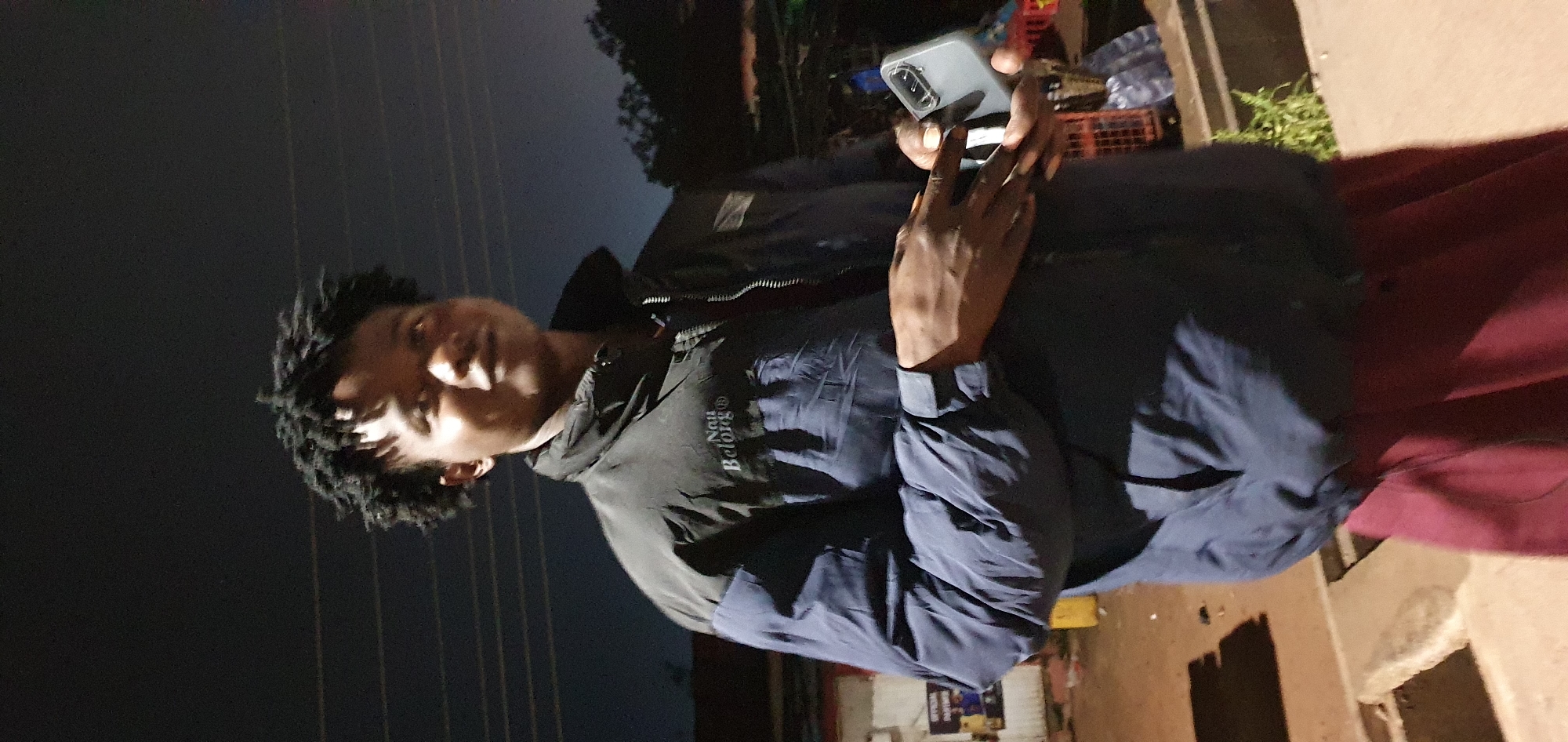 A person is standing outdoors at night in Bukuru, Nigeria, holding a smartphone. The individual is wearing a dark blue and black puffer jacket with "Nai Belong" written on the left chest, and maroon pants. The background shows a dimly lit street with some buildings and trees. The sky is dark, suggesting it is nighttime. The person's right hand is holding the smartphone, which is silver. The lighting suggests artificial lights are present, possibly from streetlights or nearby buildings.