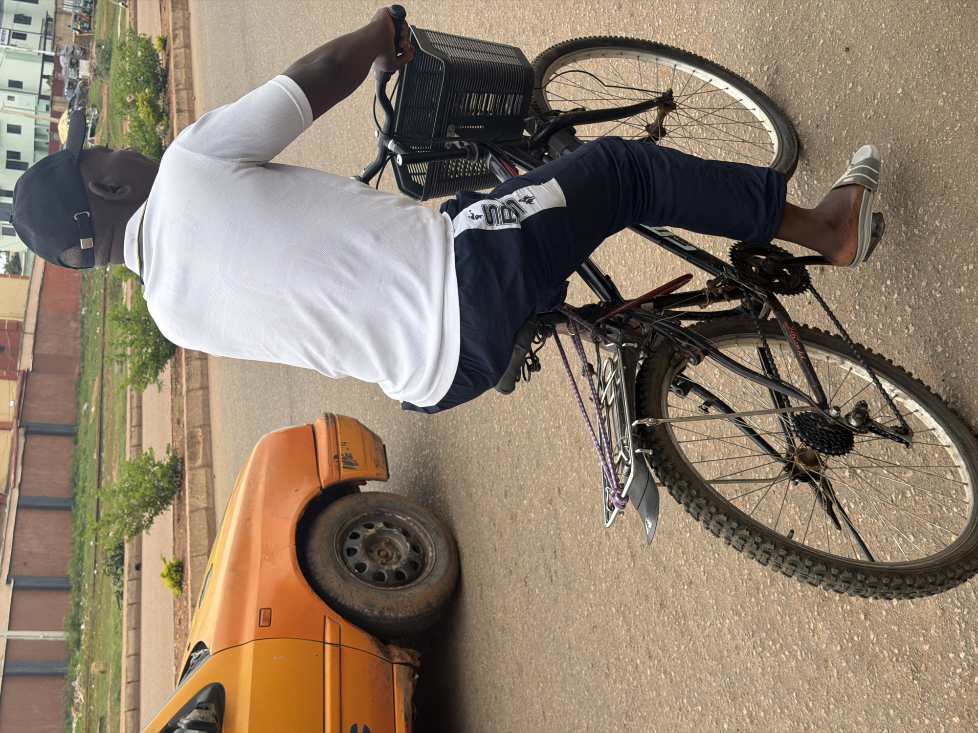 A person rides a bicycle down a paved road, seen from a low-angle, rear-facing perspective. The rider, wearing a white polo shirt, dark pants with white stripes, and a black baseball cap, is positioned on the right side of the frame, with their left leg on the pedal. The bicycle is dark-colored and equipped with a front basket and a rear rack secured with purple bungee cords.
To the left of the bicycle is the rear section of an orange car. The car appears worn, with visible rust and dirt. Its wheel is prominent in the foreground. The scene takes place on a road with a textured asphalt surface. In the background, on the left side, are buildings with multiple windows, suggesting an urban or semi-urban environment. Green foliage, including trees and bushes, lines the edge of the road.
The image is taken in daylight, with soft lighting that does not cast harsh shadows, suggesting it might be late morning or early afternoon. The overall atmosphere appears ordinary, depicting a common mode of transportation in Bukuru, Nigeria. No specific emotional cues are discernible from the rider's posture or the scene. There is no visible text in the image.