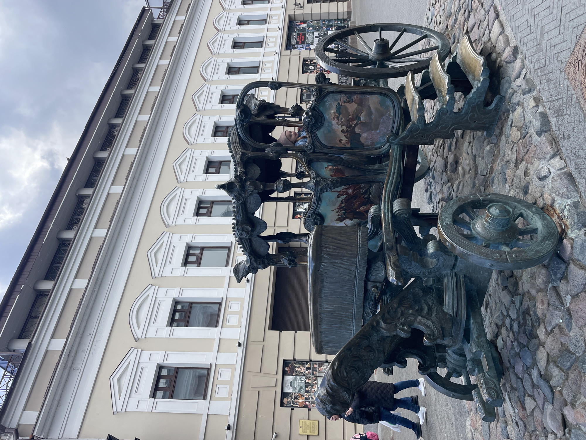This image, taken in Kazan, Russia, features a large, ornate bronze sculpture of a decorative carriage or chariot set on a cobblestone street. The sculpture is rich in detail, with intricate scrollwork, large spoked wheels, and what appear to be stylized mythical creatures, possibly lions or griffins, at its front. The interior panels of the carriage are adorned with vibrant painted or enameled scenes depicting classical or mythological figures and cherubs.
A young child, wearing a pink hat and a light blue jacket, is seated inside the sculpture, looking out. Partially visible near the base of the carriage are two adults, dressed in dark jackets and jeans, suggesting a cool, daytime environment.
The sculpture stands in front of a multi-story building with a light-colored facade, characterized by classical architectural elements including decorative moldings and windows with arched and geometric frames. The ground floor of the building features shopfronts or entrances, where posters displaying multiple faces and figures are visible, possibly advertising an event or performance. A small, dark plaque is affixed to the building wall, though its text is illegible.
The sky is overcast with grey clouds, indicating a cloudy day. The setting suggests a public square or pedestrian area in a historic city, where the elaborate sculpture serves as an artistic landmark and a point of interest for visitors.
