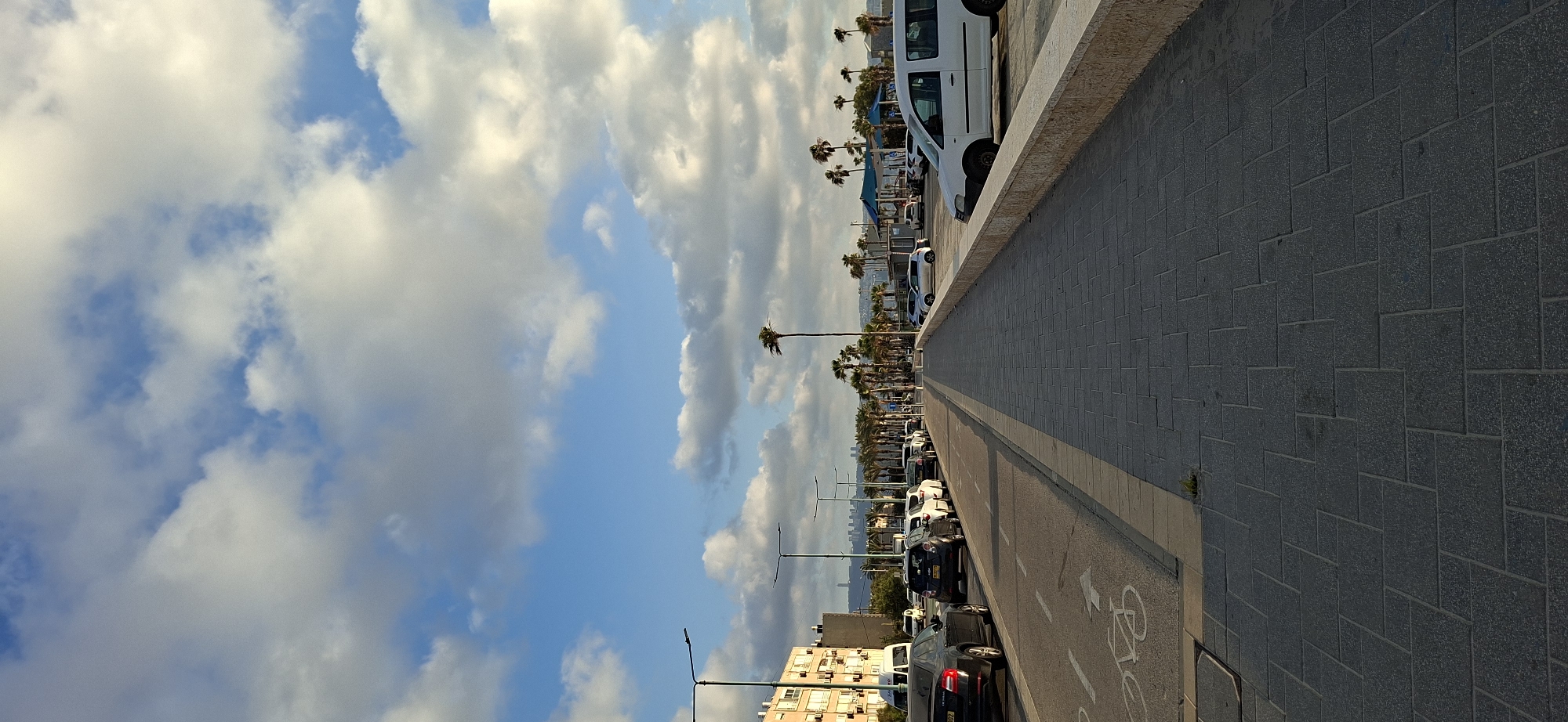 An eye-level, medium-shot, vertical, outdoor, daytime, full-body photograph captures a sidewalk extending into the distance along a coastal city street. The sky is filled with a mixture of white and gray clouds against a bright blue backdrop. On the left, a multi-story building with a tan facade stands behind a row of parked cars and streetlights. The street itself is lined with cars, and a bicycle lane is visible on the roadway. Palm trees are scattered throughout the scene, with a particularly tall palm tree standing prominently in the mid-ground. Further down the street, more palm trees line the area, and in the distance, a cityscape with buildings is visible. To the right of the sidewalk, a low stone wall separates it from a parking lot where several vehicles are parked, including a white van. The sidewalk is paved with dark gray rectangular tiles arranged in a geometric pattern.