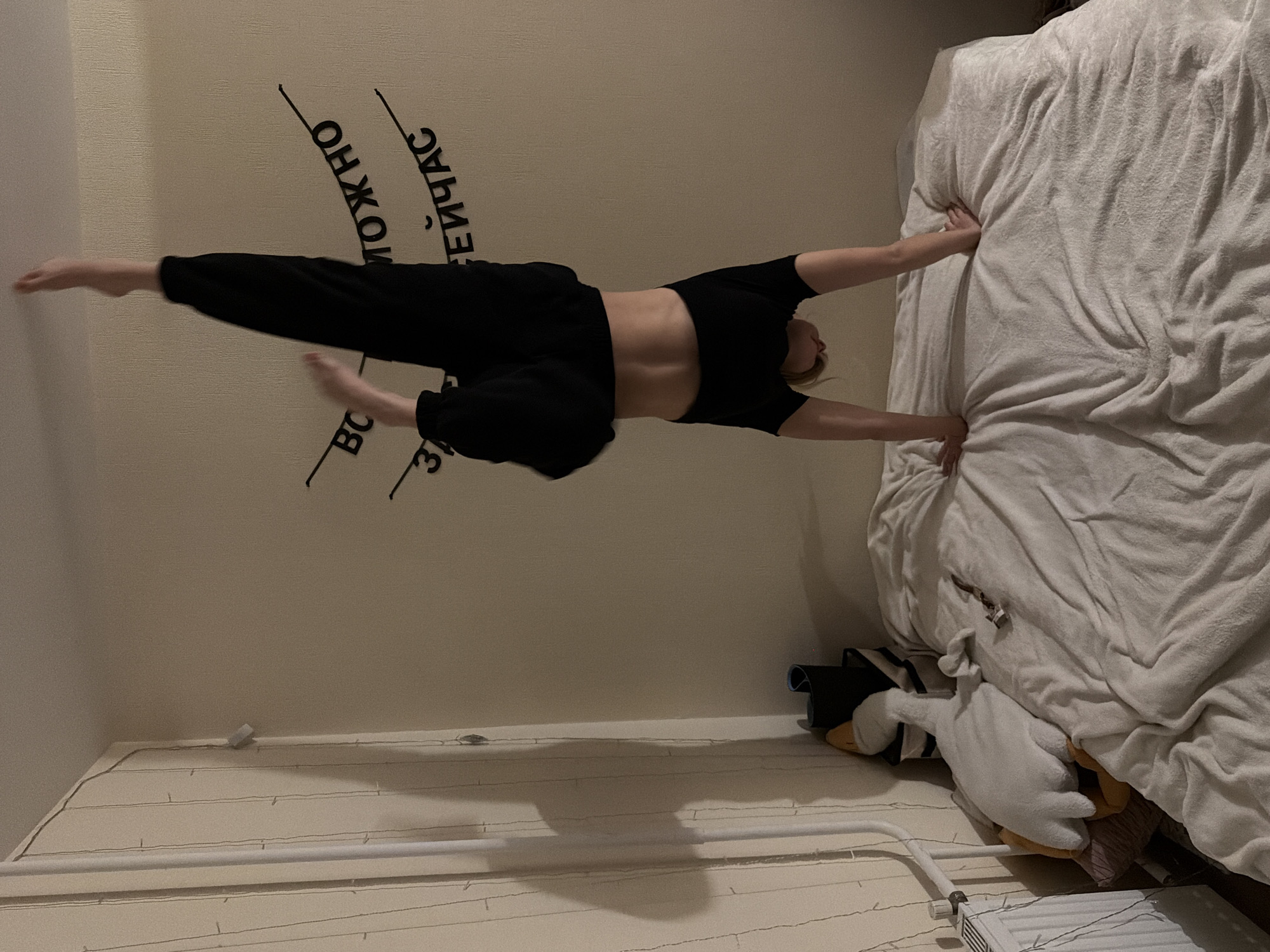 An image, rotated 90 degrees clockwise, captures a young woman performing a balancing or strength-based pose in what appears to be a bedroom in Izhevsk, Russia.
**Subjects:** The central subject is a young woman with light-colored hair, dressed in a black short-sleeved crop top and black loose-fitting pants, revealing her midriff.
**Activity or event taking place:** The woman is lying on her stomach on the floor (which appears as a wall due to the rotation). Her arms are extended forward, with her hands resting on the side of a bed, possibly for support or balance. Her legs are extended backward and lifted off the floor, creating a back extension or "Superman" pose commonly seen in yoga or pilates. Her feet are pointed. She appears to be focused on maintaining the pose.
**Scene and Setting:** The setting is an indoor room, likely a bedroom. The floor (appearing as a wall) is a light, possibly beige, color and features decorative black text. String lights are laid out along the bottom of the floor/wall. A bed with rumpled white or light-colored bedding is positioned against a wall (appearing as the floor).
**Notable Details:**
* **Perspective/Rotation:** The image is rotated, making the floor appear as a vertical wall and the bed as a horizontal surface. This gives the illusion that the woman is defying gravity, but she is actually prone on the floor.
* **Objects:** Besides the woman and the bed, a white radiator is visible along the actual bottom wall. A white stuffed goose plushie and a rolled-up black yoga mat are seen near the base of the bed.
* **Lighting:** The lighting is artificial, suggesting it might be evening or nighttime indoors.
* **Visible Text:** On the floor, several Cyrillic words are visible in black lettering. The discernible parts read "МОЖНО" (MOZHNO), "СЕЙЧАС" (SEICHAS), and "ВС" (VS), likely part of "ВСЁ" (VSYO). Together, they form "ВСЁ МОЖНО СЕЙЧАС," which translates to "Everything is possible now."