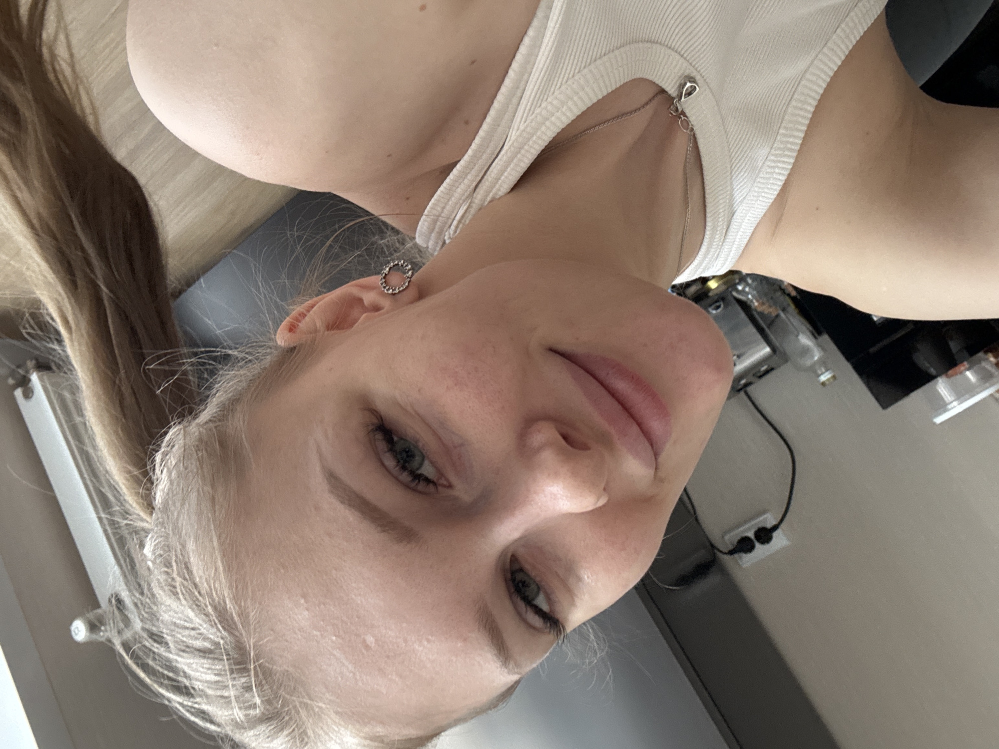 A fair-skinned woman with blond hair in a ponytail is indoors. She wears a white tank top and a circular earring. The background shows kitchen elements: a counter, appliances, and a wall outlet. There is no indication of Perm, Russia, in the image.