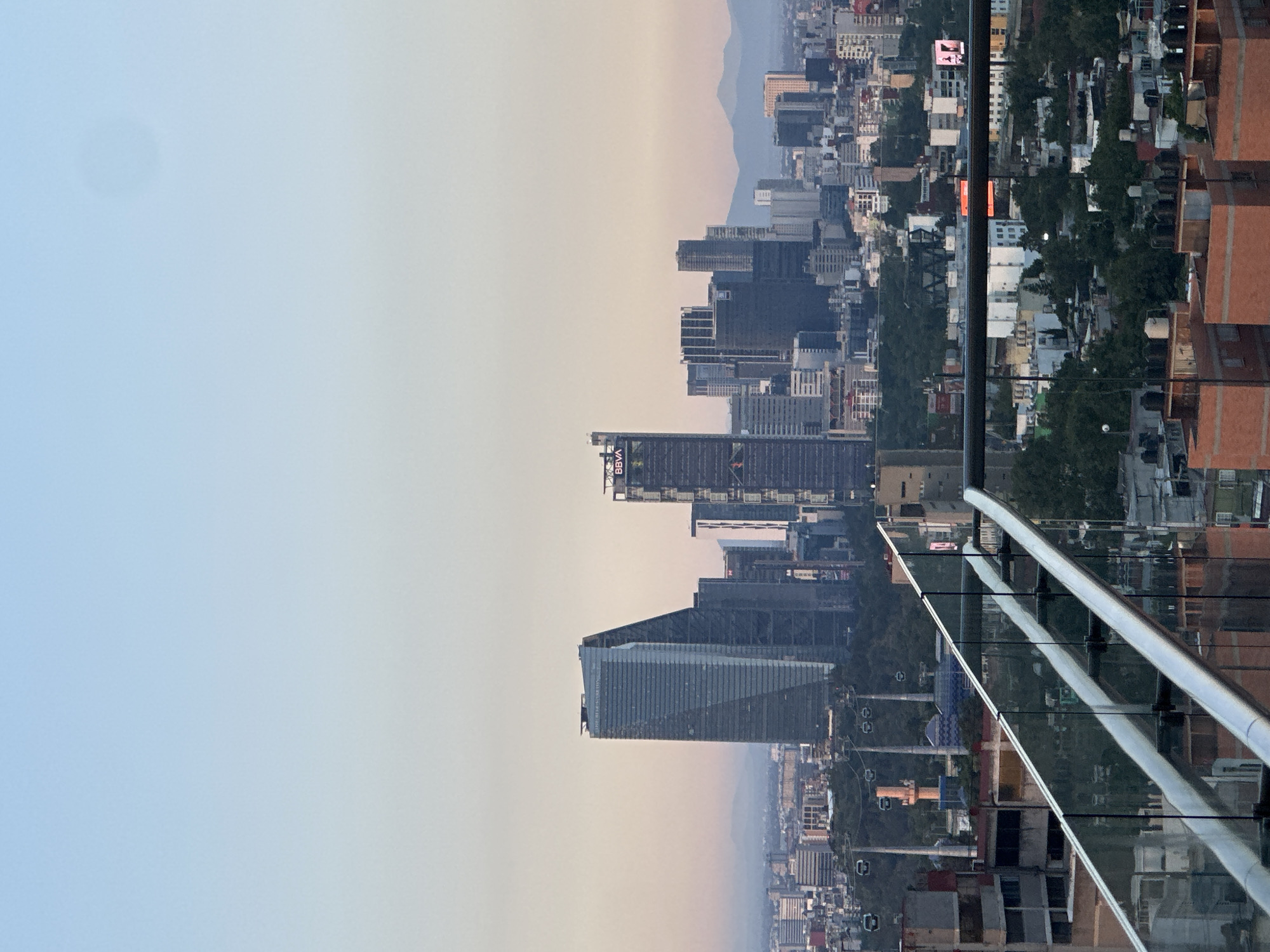 The image captures a cityscape in Mexico City, Mexico, with a prominent view of high-rise buildings. A metal and glass railing is in the foreground, suggesting the photo was taken from a high vantage point, likely a building's observation deck or balcony. The sky is a gradient of light blue to pale orange, suggesting either sunrise or sunset. The urban landscape features a mix of architectural styles, from modern skyscrapers to more traditional low-rise buildings. The background includes a faint outline of distant mountains, adding depth to the scene. A cable car system appears to be running through the city. There are no people visible in the frame.