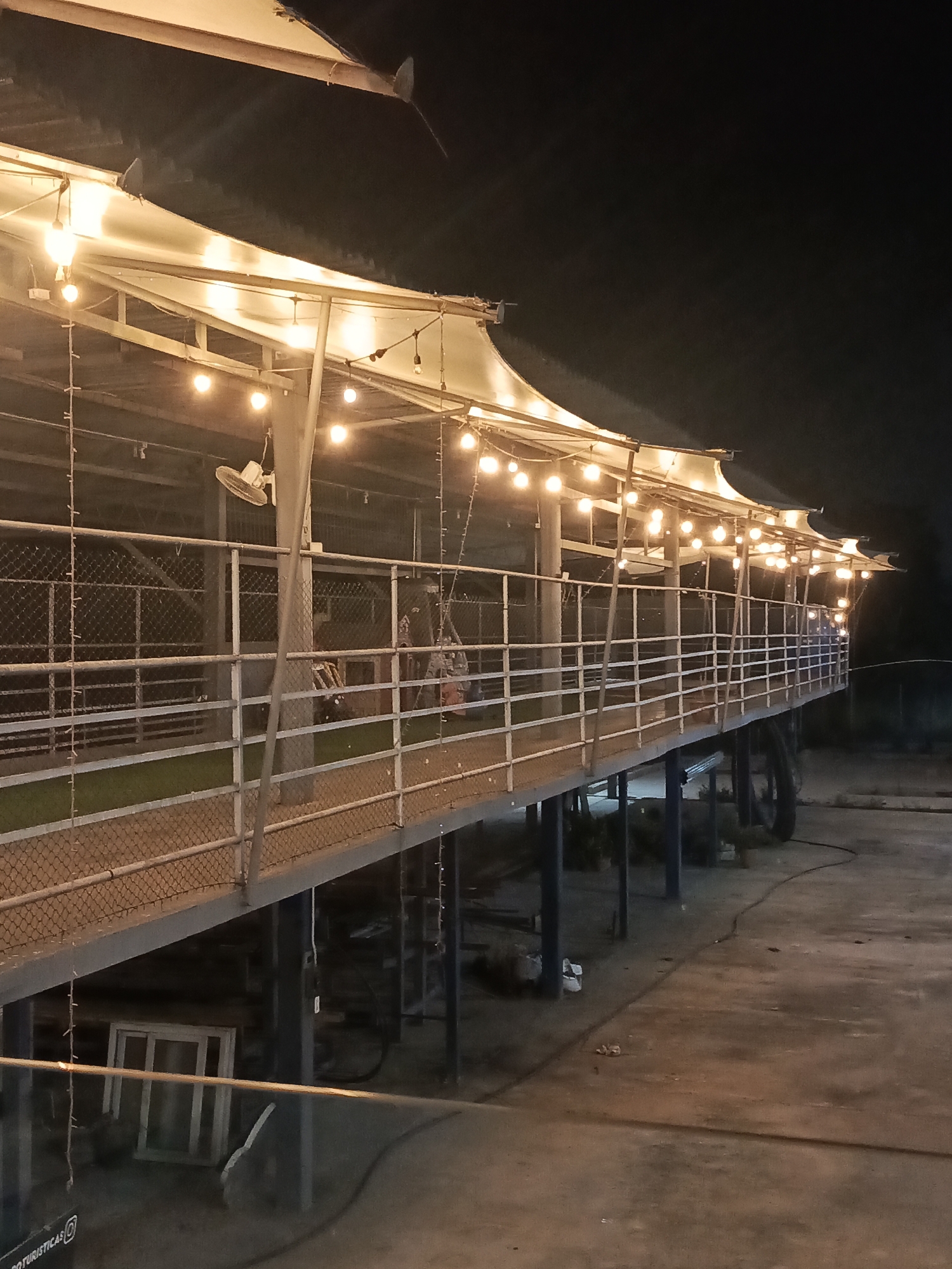 The image captures an elevated outdoor walkway or structure at night, illuminated by numerous warm-toned string lights. A white, tent-like canopy covers the walkway, reflecting the light. The structure features a metal railing with a chain-link fence running along its length, and green artificial turf is visible on the walkway's surface. A white ceiling fan hangs from the canopy structure. Hanging vertically along the railing are delicate fairy lights, adding to the festive ambiance.
Below the elevated platform, dark support beams are visible, along with a concrete ground. Items like a white window frame, a large tire or wheel, and other indeterminate objects are stored or left on the ground beneath the structure. The background is dark, indicating a nighttime setting with no visible natural light. No people are present, suggesting the area might be closed or temporarily unoccupied.
The overall atmosphere is warm and inviting due to the soft lighting, hinting at a space designed for social gatherings or dining. The scene is located in Tocuyito, Venezuela. In the bottom left corner of the original image, faint text reads "@FUTURISTICALO".