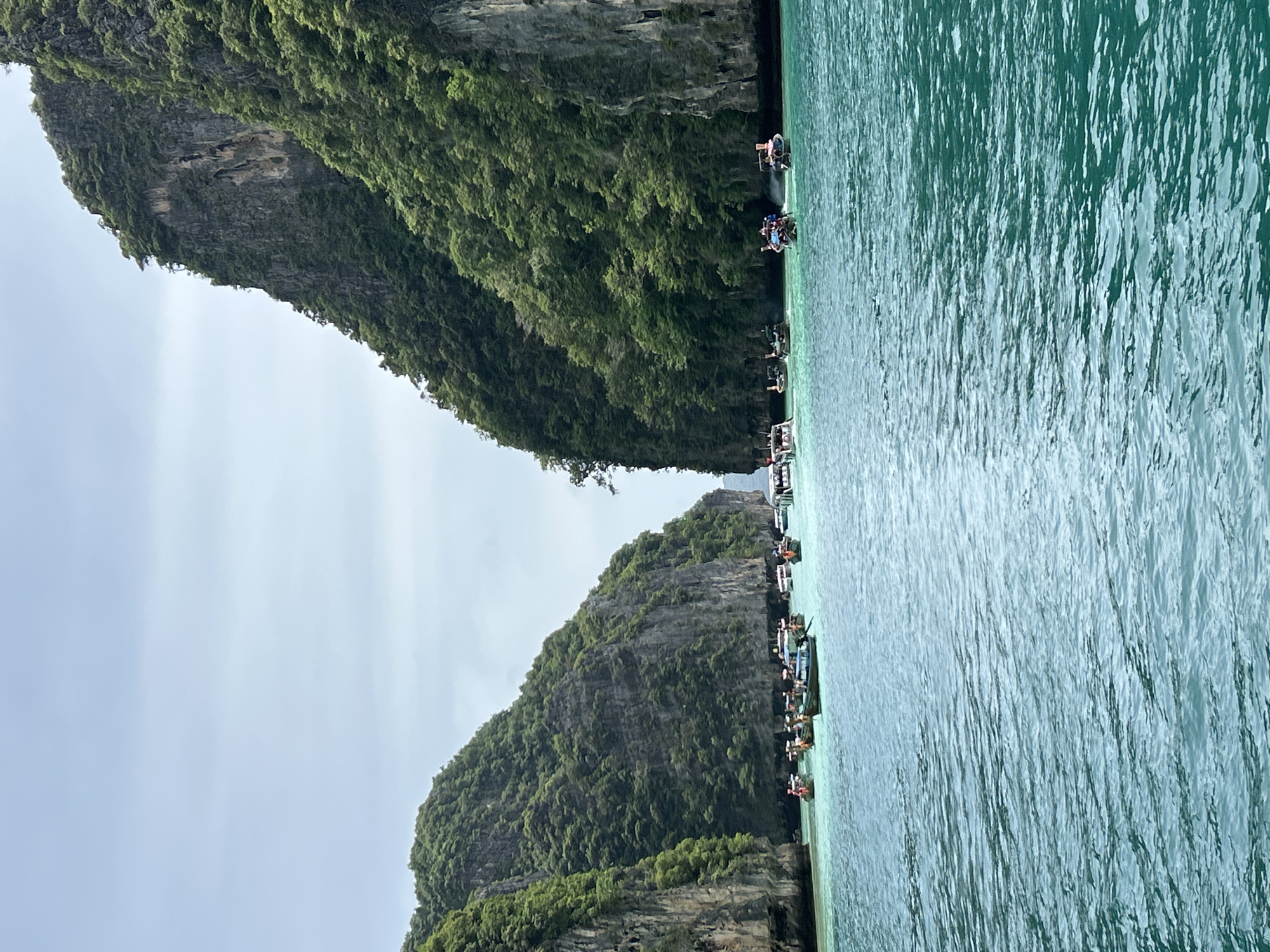 The image depicts a wide shot of a channel between two steep, lush green limestone cliffs, characteristic of the Phi Phi Islands in Krabi Province, Thailand, near Ao Nang. The water is a clear, turquoise hue, with small ripples indicating a calm sea. Numerous long-tail boats, a common mode of transport in the region, are scattered across the water. Some boats are anchored, while others appear to be in motion, carrying passengers. The cliffs are densely vegetated with tropical trees and vegetation, their rocky faces visible in places. The sky above is overcast, casting a diffused light on the scene. There are no discernible individuals in the foreground, but people are visible on the boats in the middle ground. The overall impression is a popular tourist destination offering scenic boat tours.