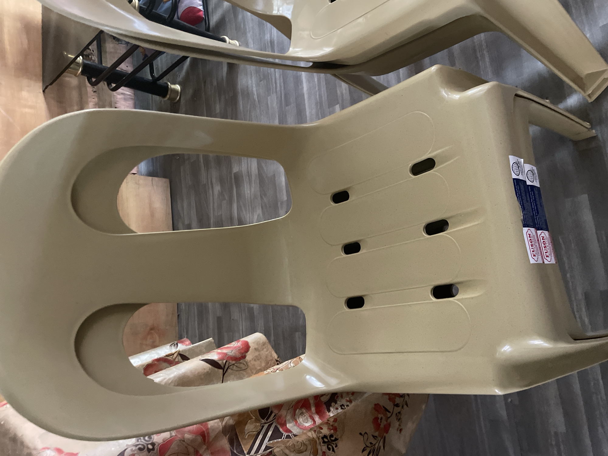 This forensic description pertains to an interior scene located in Davao City, Philippines. The central focus is a beige, injection-molded plastic chair, presented facing away from the viewer. The chair's backrest features two prominent, symmetrical arched cutouts. The seat is equipped with six oval-shaped perforations, arranged in two rows of three. Affixed to the forward edge of the seat are two white rectangular labels, each displaying the brand "FUSON" in red text, accompanied by smaller, illegible product information and circular logos.
To the right of the primary chair, a second identical beige plastic chair is partially visible, showing its backrest and a portion of its seat. On the left side of the frame, a multi-colored floral tablecloth, patterned with large red roses, green foliage, and brown/beige abstract designs on a light background, is draped over an unseen object, likely another chair or table. The floor is covered with grey, wood-grain laminate planks. In the background, through the cutouts of the main chair's back, a light-toned wooden surface is discernible, alongside a dark metal table leg accented with gold-colored rings, indicating the presence of a table or similar furniture. No individuals or active subjects are present in the image.
