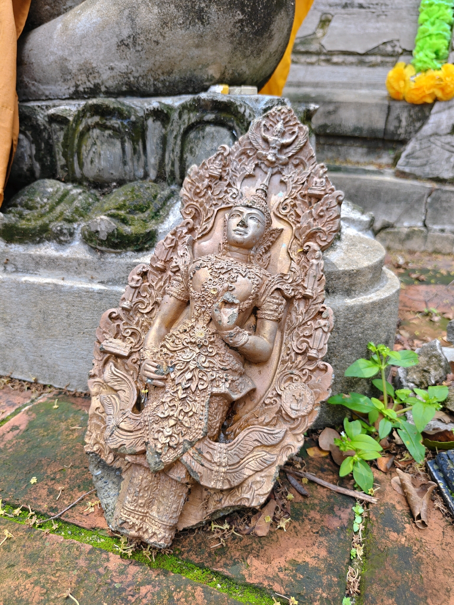 A broken stone statue of a deity, possibly a depiction of a celestial being or goddess, lies on a weathered brick surface in Phra Nakhon Si Ayutthaya City Municipality, Thailand. The statue, rendered in a metallic bronze color, is intricately carved with ornate details, including elaborate headdresses, layered clothing, and decorative patterns. It appears to be a fragment, with the lower portion missing or broken away. The statue rests on a reddish-brown brick ground, which is partially covered by green moss and scattered fallen leaves and twigs. In the background, weathered concrete structures, possibly part of a temple or ancient ruins, are visible. A portion of a larger, light-colored stone structure, perhaps another statue or architectural element, is partially obscured by an orange fabric draped over it. The environment suggests an outdoor setting within a historical or religious site, possibly exposed to the elements, leading to the statue's current damaged state.