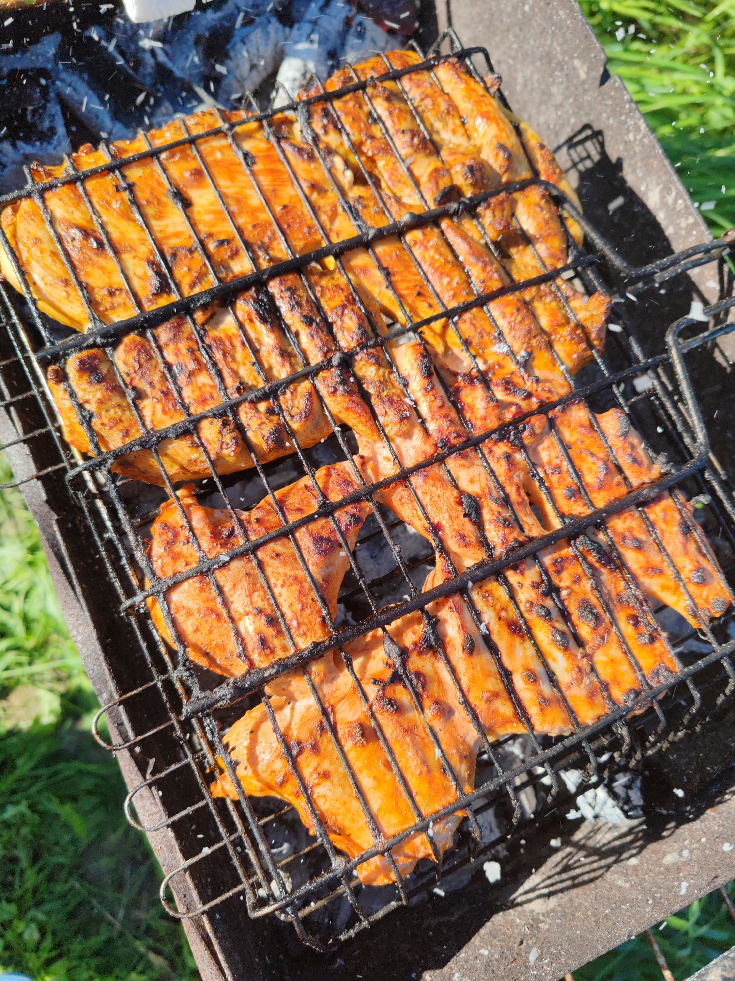 In Чухлэм, Russia, chicken pieces are grilling on a black metal barbecue rack. The chicken appears to be coated in a red-orange spice blend, and some pieces show char marks from the heat. The barbecue is placed outdoors, with green grass visible in the background, suggesting a sunny day and a potential picnic or outdoor gathering. Ash from the coals is scattered around the grill, and there are hints of white ash falling from above, possibly from the burning charcoal or wood. The scene is captured from a high-angle, looking down at the cooking food.