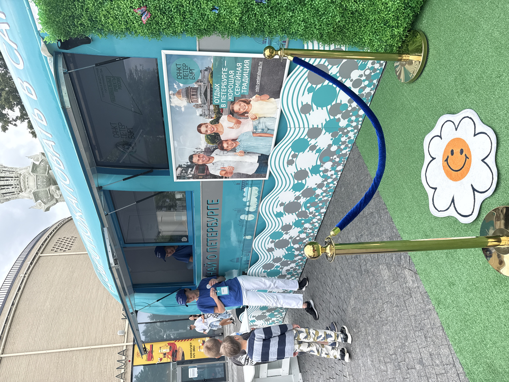 This is a medium-shot, eye-level photograph taken outdoors on a cloudy day. In the center, a bright teal food truck with the words "ДОБРО ПОЖАЛОВАТЬ В САНКТ-ПЕТЕРБУРГ" (Welcome to Saint Petersburg) written in white lettering across its top. The truck has a serving window open, and two individuals wearing blue shirts and dark caps are visible inside. A woman in a blue t-shirt and white pants stands in front of the truck, facing three young boys. She appears to be interacting with them.
To the left of the truck, part of a building with "LAMA" visible on its facade can be seen. On the right side of the truck, a tall, artificial green hedge with two pink butterflies on it stands. In front of the truck, a gold stanchion with a blue velvet rope guides visitors. On the grass in front of the stanchion is a white and orange flower-shaped mat with a smiley face. The ground is paved with grey cobblestones in the background and artificial green turf in the foreground, indicating an outdoor event or exhibition. The overall atmosphere is cheerful and inviting. Based on the context of promotional materials for St. Petersburg, this scene could plausibly be from an event in Moscow, Russia.