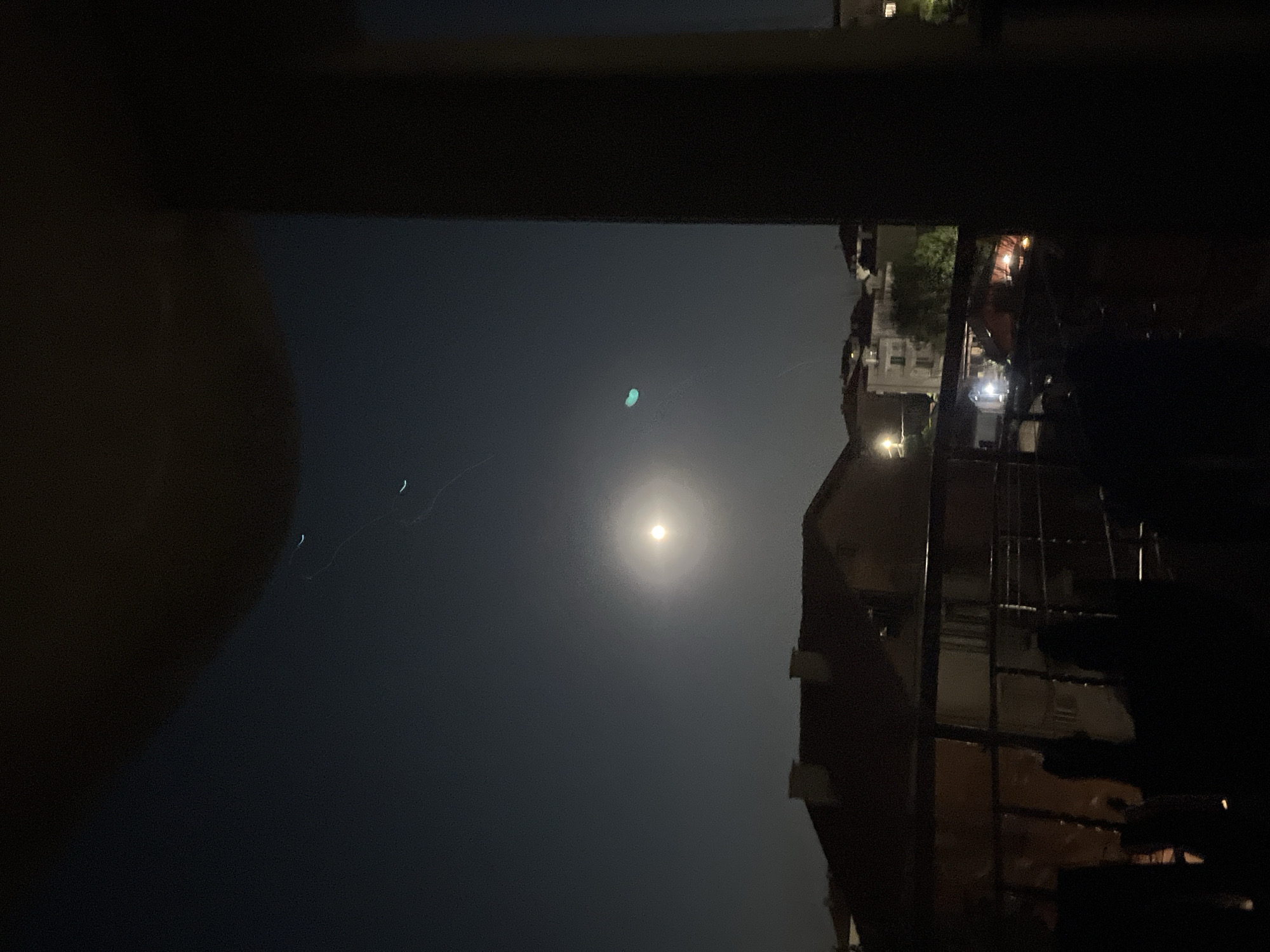 The image depicts a night scene from a balcony overlooking a residential area in Obzor, Bulgaria. A full moon is visible in the dark sky, surrounded by a bright halo. Several faint, curved light trails are scattered across the sky, possibly from a drone or aircraft. Below the sky, rooftops of houses and apartment buildings are visible, illuminated by streetlights and interior lights. The foreground shows the dark silhouette of a balcony railing and parts of the balcony itself. The overall impression is a quiet, urban night.