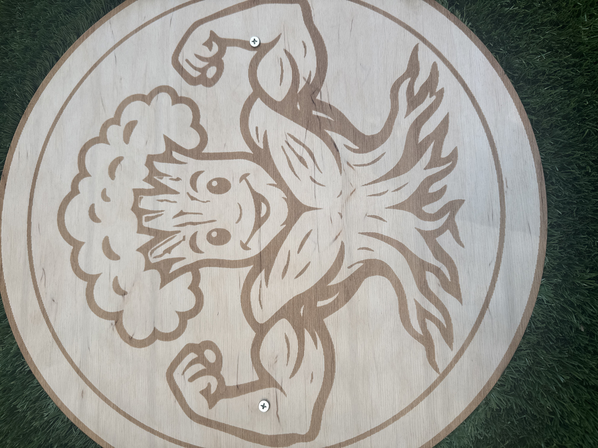 A close-up, overhead shot shows a circular wooden plaque with a carved image of a muscular cartoon tree character with its arms flexed. The character has a smiling face with large eyes and a cloud-like crown of leaves. The wood has a visible grain pattern and is secured to a surface by two visible silver screws. The plaque rests on a patch of artificial green turf. The image is centrally framed, with a portion of the turf visible at the top and bottom edges of the frame. The plaque is positioned on the ground, suggesting an outdoor or garden setting. No specific location in Sofia, Bulgaria is identifiable from the image.