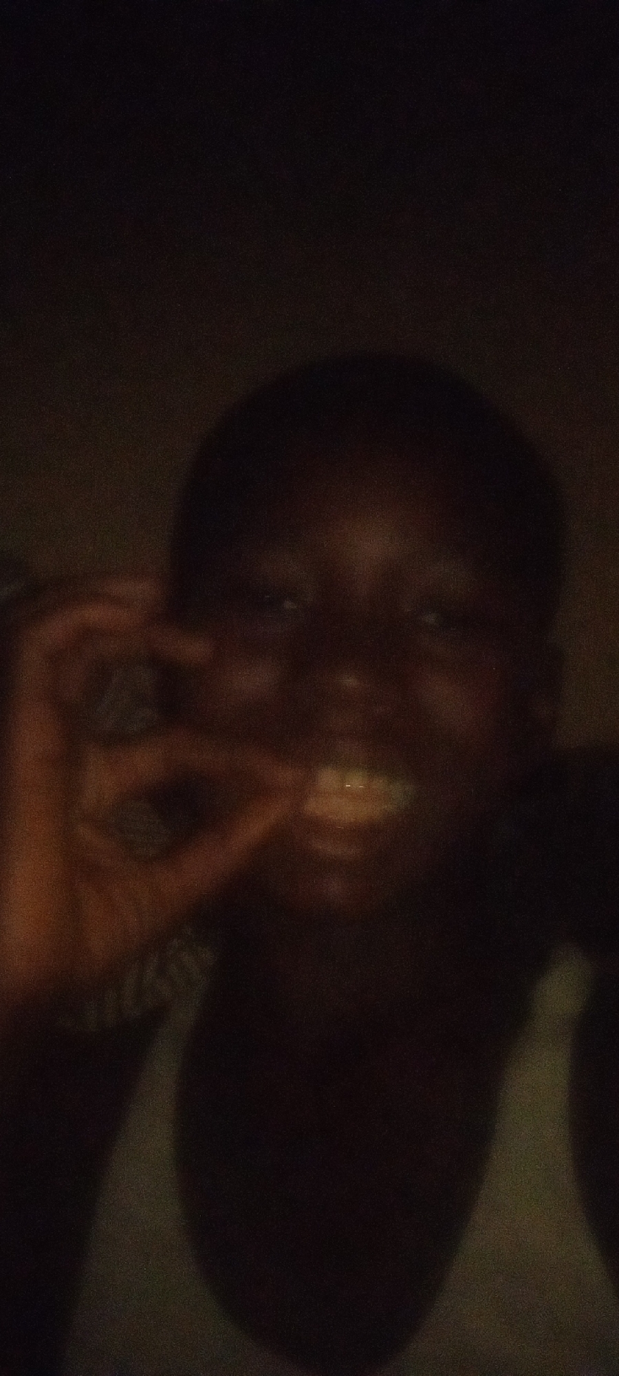 A dark, blurry image appears to show a person with dark skin and short hair. The person is wearing a light-colored sleeveless top. Their hand is raised near their face in a gesture that is difficult to discern due to the poor image quality. Their mouth is open, possibly smiling. The background is dark and indistinct. The location in Adankolo, Nigeria is not apparent from the image.