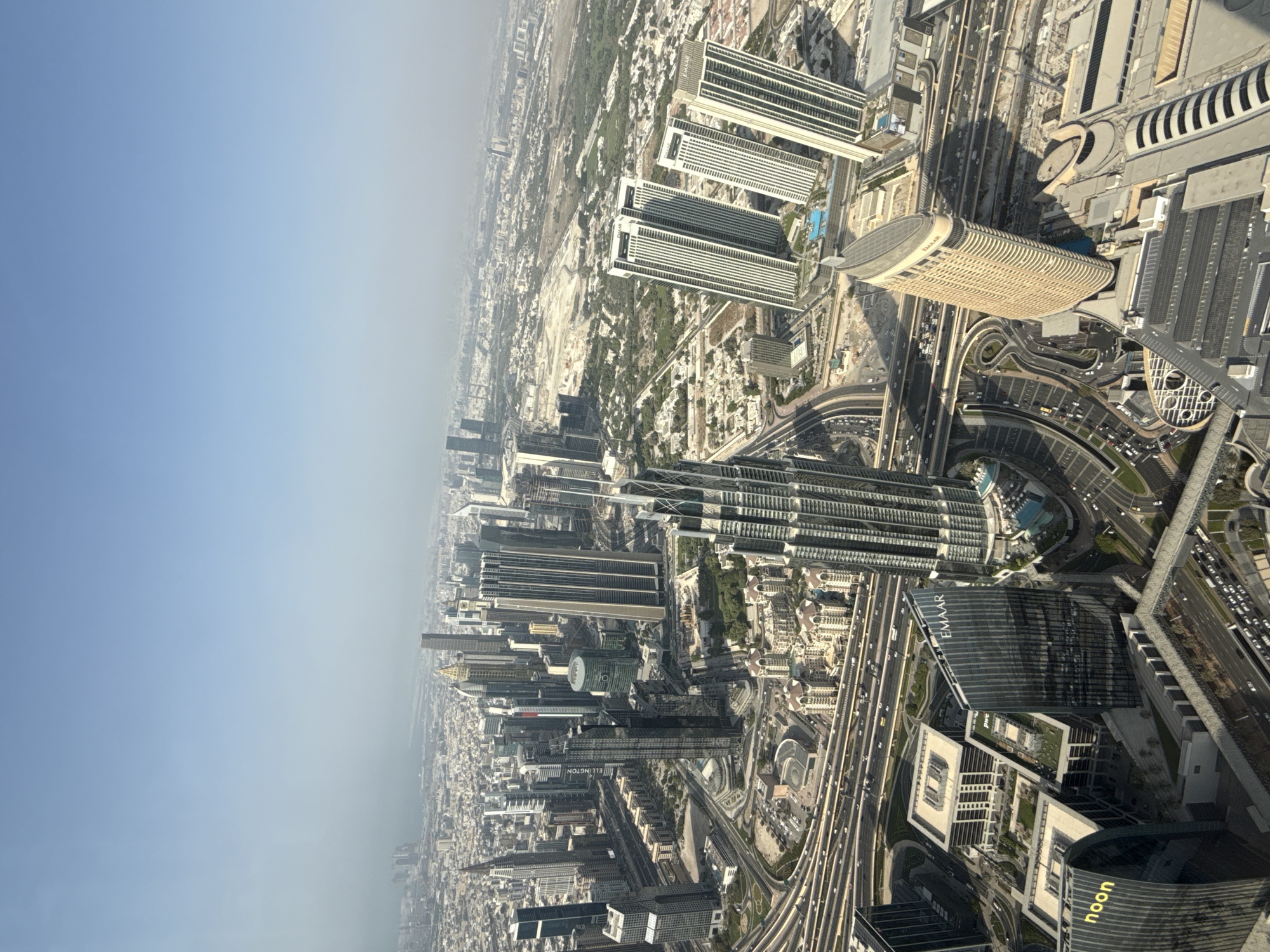 This is a high-angle, wide-shot aerial view of Dubai, United Arab Emirates, likely taken from a very tall skyscraper. The image captures a dense urban landscape dominated by modern architecture, including numerous skyscrapers of varying heights and designs. The iconic Burj Khalifa is visible in the mid-ground, its distinct silhouette piercing the hazy sky. Surrounding it are other notable buildings such as the Address Downtown Dubai and the Emaar headquarters. A complex network of multi-lane highways and elevated roads, characteristic of Dubai's infrastructure, snakes through the cityscape, with traffic visible on some segments. Low-rise buildings, residential areas, and green spaces, including parks and possibly golf courses, are also interspersed throughout the broader urban expanse, extending towards the horizon where a faint, hazy outline suggests the presence of the sea or a distant landmass. The lighting suggests it is daytime, with the sun casting shadows from the buildings. The sky is a pale blue, indicating a possibly warm and humid atmosphere, with a haze obscuring the far distance. No people are discernible in the image.