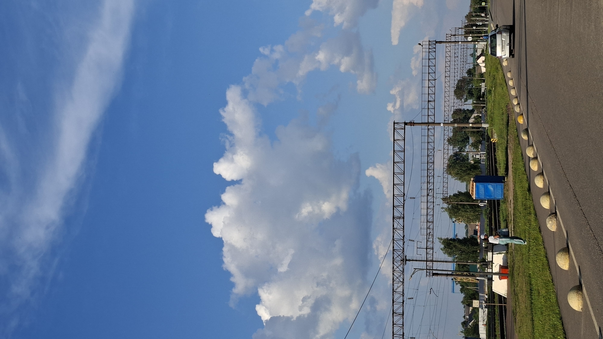 The image shows a street scene in Smolevichi, Belarus. The sky is a vibrant blue with scattered white clouds. In the foreground, a paved road curves to the right, lined with spherical concrete bollards. To the left of the road is a grassy verge. Beyond the verge are railway tracks with overhead electrical lines supported by metal structures. A blue portable toilet is visible near the tracks, and a person wearing a white hat, light blue shirt, and blue pants stands nearby, holding a white bag. Further in the background, there are trees, buildings, and a silver car driving away on the road. The scene appears to be a semi-urban or industrial area.
