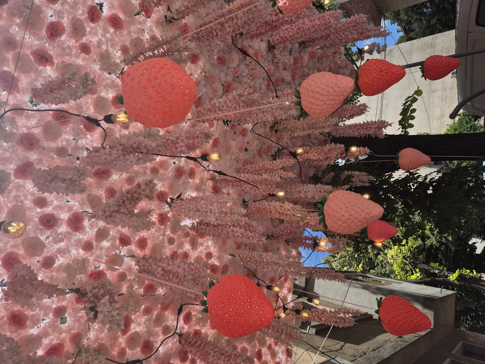 An upward angle shot of a floral installation with large strawberry decorations and string lights, suggesting an outdoor event or decorated public space. The canopy is densely covered with artificial pink and white roses, interspersed with cascading pink wisteria-like flowers. Several large strawberry-shaped ornaments, some red and some pink, hang from the floral canopy by pink strings. Incandescent string lights are also visible, adding to the festive atmosphere. Below the canopy, glimpses of a building facade, green trees, and a clear blue sky are visible. The scene appears to be in an urban environment, possibly a street or courtyard, given the presence of a building and what looks like a vehicle at the bottom right. There are no people in the image.