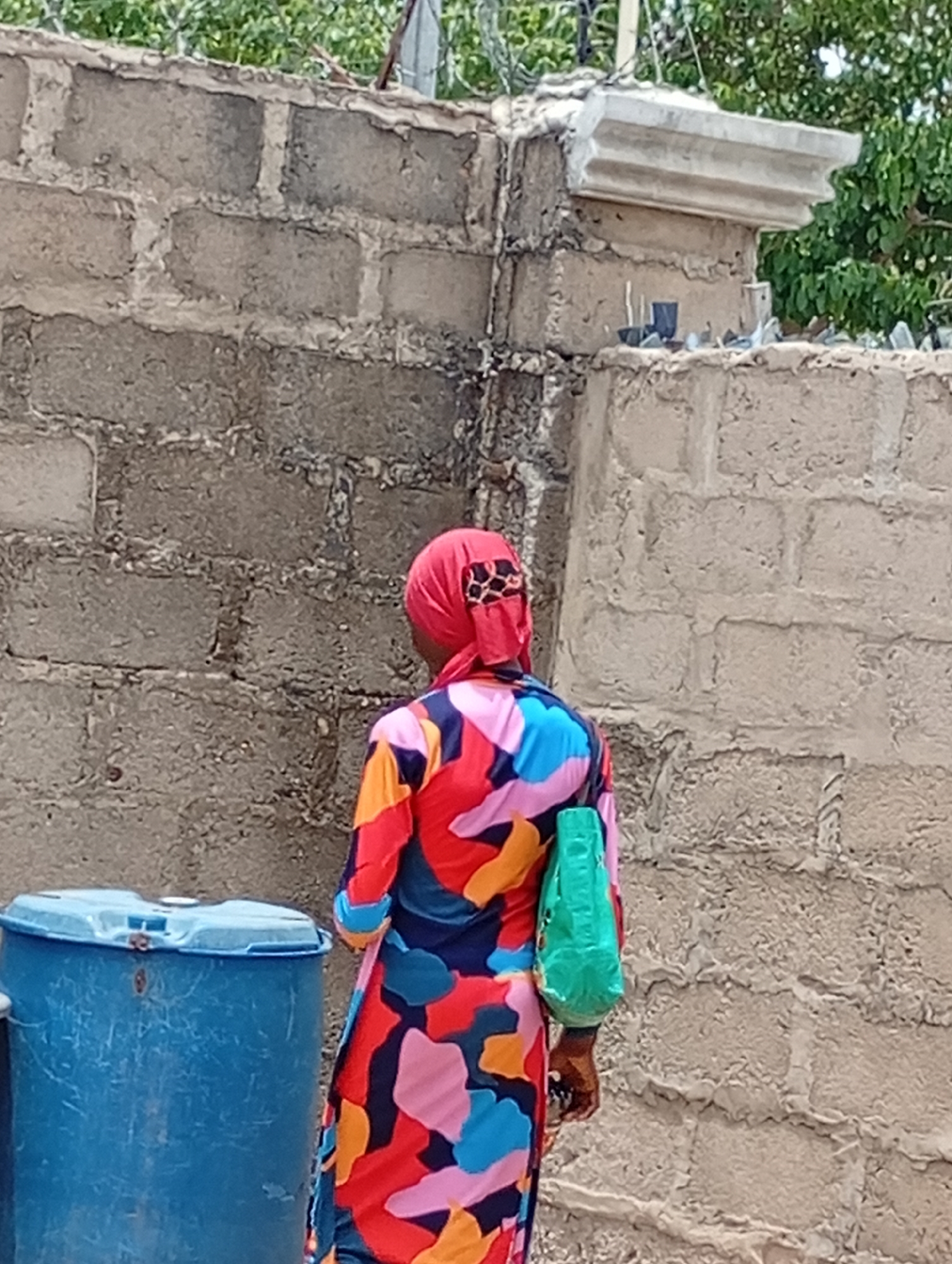A woman with a red headscarf and a colorful, patterned dress stands next to a blue plastic barrel in front of a grey concrete block wall. The woman is facing away from the camera and carrying a green bag. The wall is topped with barbed wire and broken glass, likely for security. The image appears to be taken in an outdoor urban environment, possibly in the city of Lokoja, Nigeria.