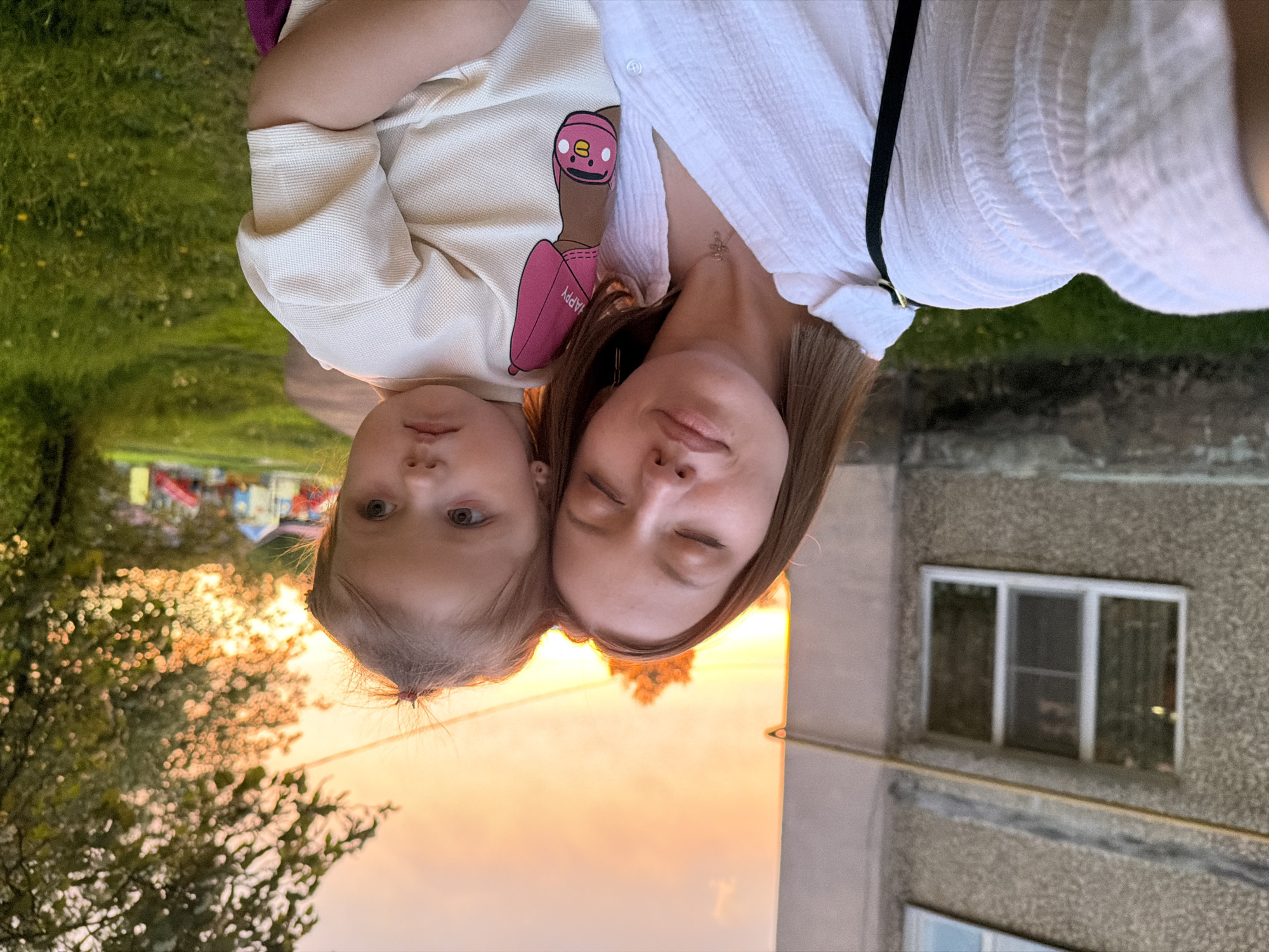 A selfie photograph shows a fair-skinned woman and a child. The woman has long brown hair and is wearing a white shirt. She is holding the child, who is wearing a light-colored shirt with a cartoon graphic.
The background shows elements of an urban environment, including a multi-story concrete building with a window, trees, a grassy area, and a playground visible in the distance. The sky has a warm, sunset glow.
Based on the location, this is likely a residential area in Reshetnikovo, Russia.