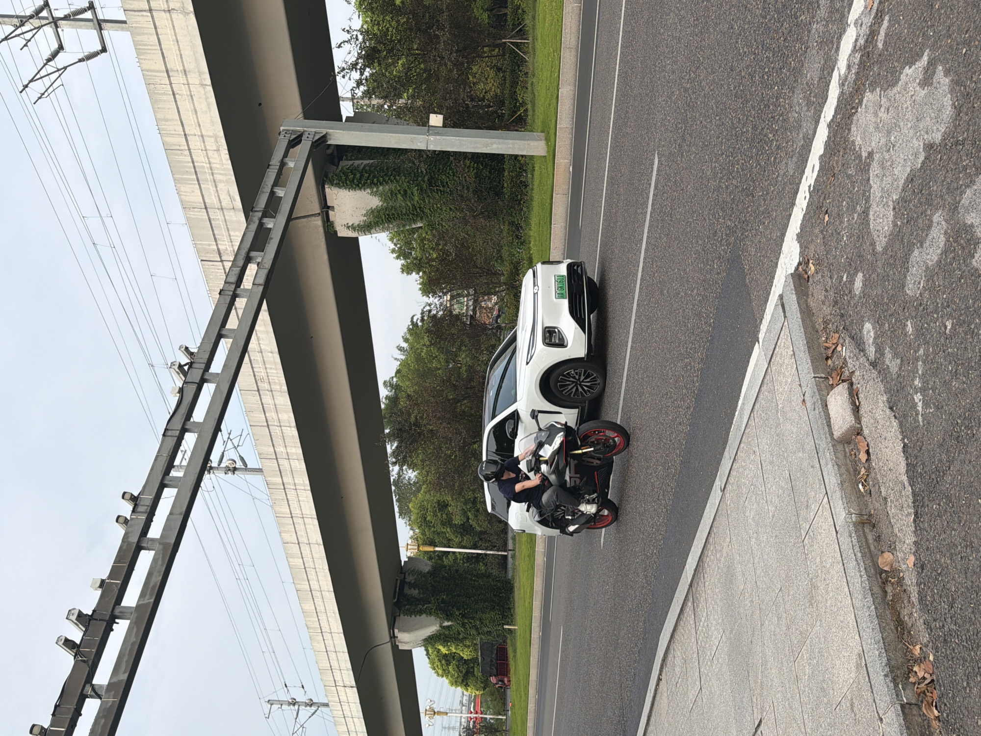 In the city of Jiangjin, China, a motorcycle rider wearing a black helmet and a dark blue t-shirt is operating a black and red sport motorcycle on a paved road. The motorcycle has red rims on its wheels. The rider is positioned in the left lane of the road. To the right of the motorcycle, a white SUV is traveling in the same direction, also in the left lane. The SUV has a green license plate with the characters "A GKI611". Both vehicles are moving under a large, elevated concrete structure, which appears to be an overpass or bridge for a railway line. This structure is supported by concrete pillars that are partially covered in green ivy. Overhead, power lines and what appear to be security cameras are mounted on the elevated structure. The road is bordered on the left by a sidewalk made of gray paving stones. The background consists of lush green trees and vegetation, and the sky is overcast.