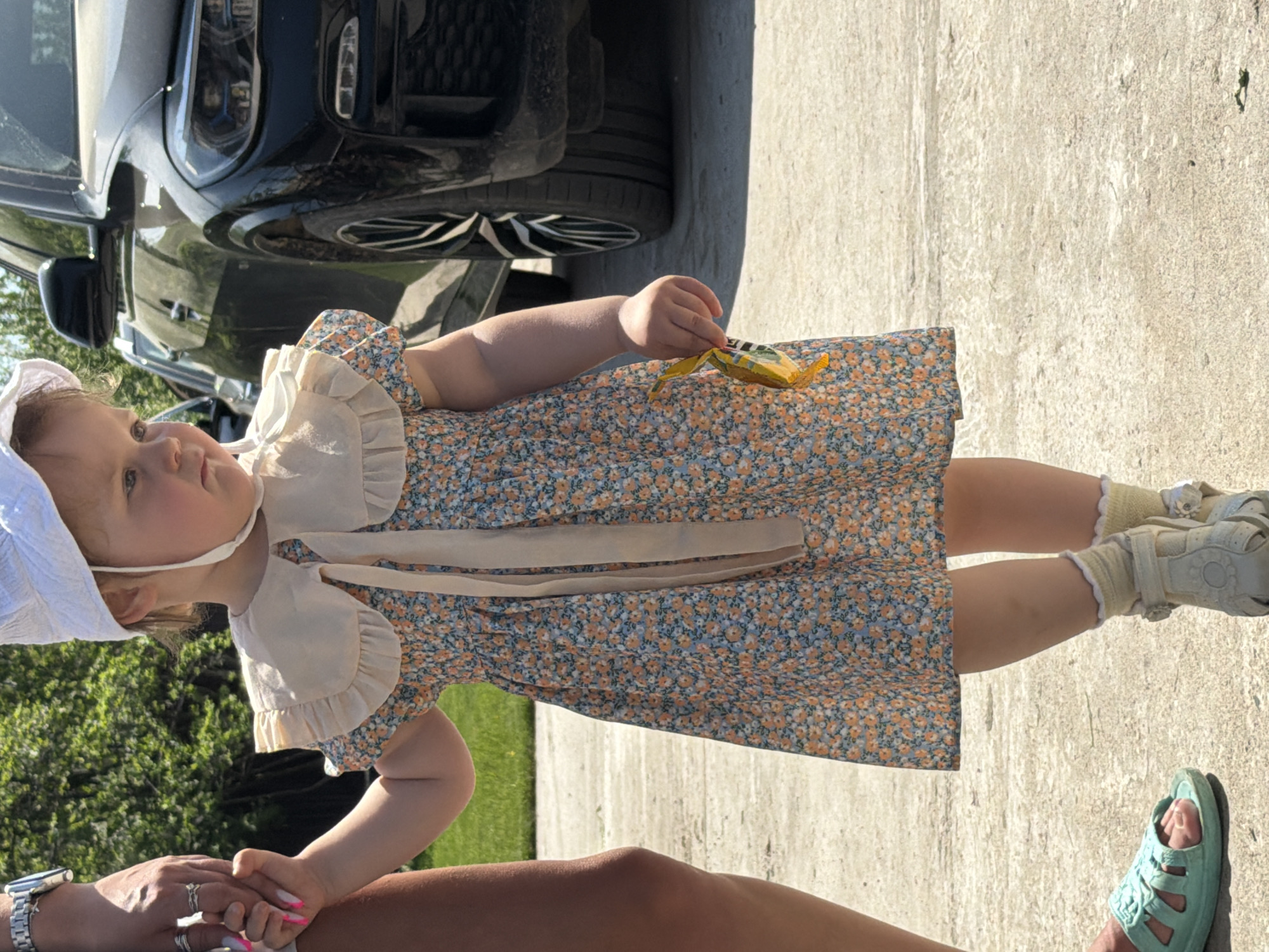 The image depicts a child in a blue and orange floral dress and a white bonnet. The child holds a yellow object in her right hand. She is wearing light-colored socks and shoes. A car is visible in the upper left portion of the image. The child appears to be standing on a paved surface. A person wearing a watch is holding the child's hand. Another person is wearing a teal sandal. The setting appears to be outdoors, with some green vegetation visible in the background. There is no information to support the image being in the city of Chaikovsky, Russia.