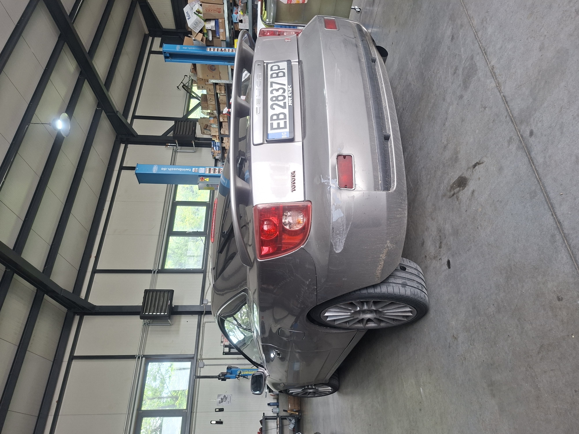 The image shows the rear of a gray Toyota Celica parked inside a large garage or workshop. The vehicle has a Bulgarian license plate that reads "EB 2837 BP." The car appears slightly dirty and has some scratches or imperfections on its rear bumper. The garage has a high ceiling with exposed metal beams. There are windows along the walls, and a blue vehicle lift is visible in the background. There are also boxes and equipment in the background. The floor of the garage is concrete and appears to be slightly stained. No people are visible in the image. The location in the city of Unknown, Unknown is deduced from the license plate being Bulgarian.