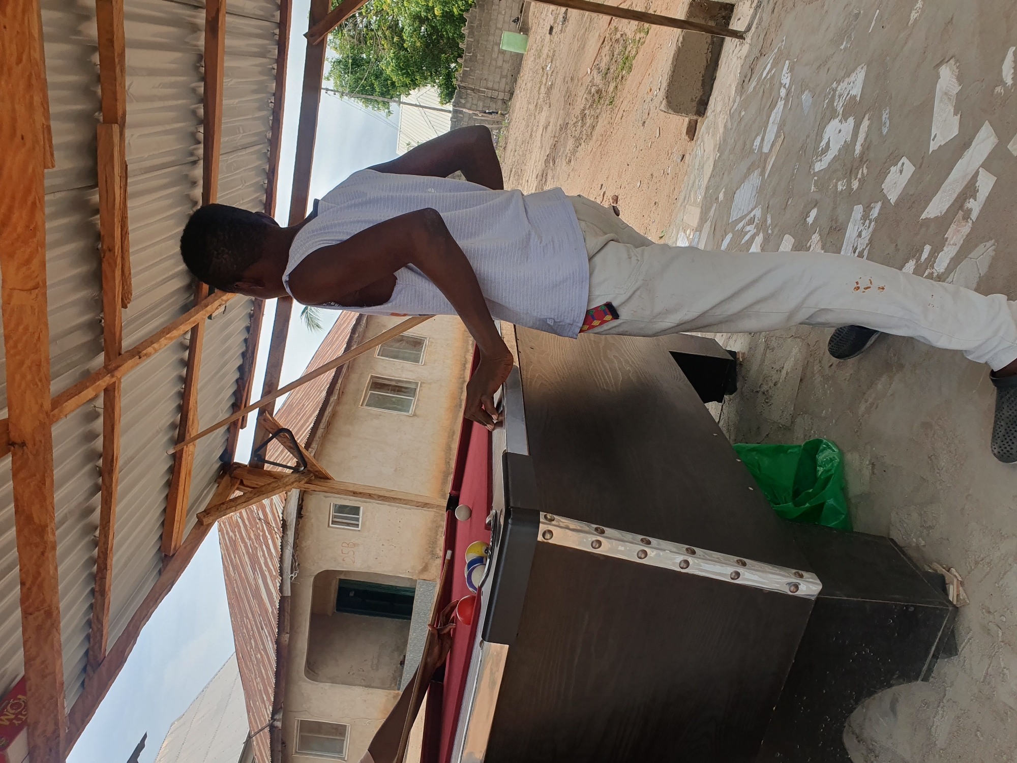 The image depicts an outdoor scene, likely in a semi-urban or rural area of Lokoja, Nigeria. A young Black male, wearing a light blue and white striped tank top and light-colored pants, stands near a billiard table. He is facing away from the camera, looking towards the billiard table. Another individual's hands are visible on the billiard table, holding a cue stick, suggesting a game of billiards is in progress or about to begin. The billiard table itself is dark-colored with a red felt surface and appears to be well-used. Several billiard balls are on the table, including a red ball, a striped ball, and a white cue ball.
The environment surrounding the billiard table suggests a rustic setting. A corrugated metal roof supported by wooden beams covers the area, providing shade. In the background, a simple, light-colored building with a corrugated metal roof is visible. The ground is a mix of sand and concrete, with some debris and a green plastic bag on the ground near the billiard table. A stone wall and some trees are visible further in the background, indicating a potentially dry or seasonal climate. The overall impression is a casual, communal gathering space for recreation.