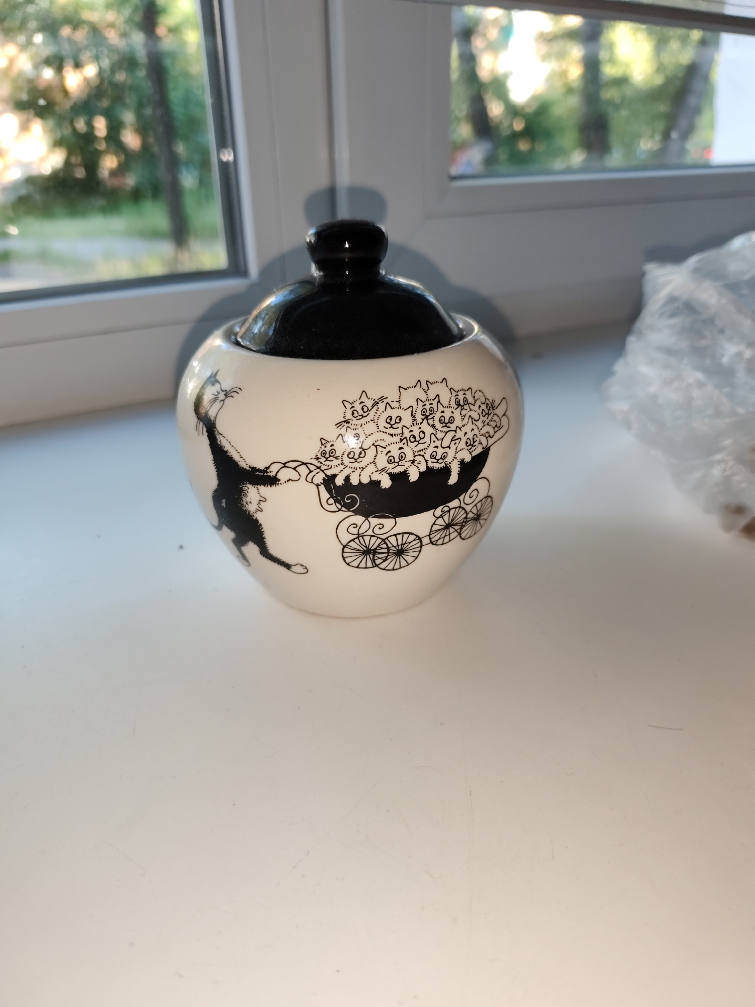 A black-lidded, white ceramic sugar bowl with a black drawing of cats is placed on a white surface. The drawing depicts a cat pulling a carriage filled with multiple kittens. The sugar bowl is situated near a window that offers a view of green trees and sunlight. A crumpled clear plastic bag is visible to the right of the sugar bowl. The scene is indoors, with natural light entering through the window. The objects are positioned on a windowsill or a similar surface in a home environment. There are no people present in the image.