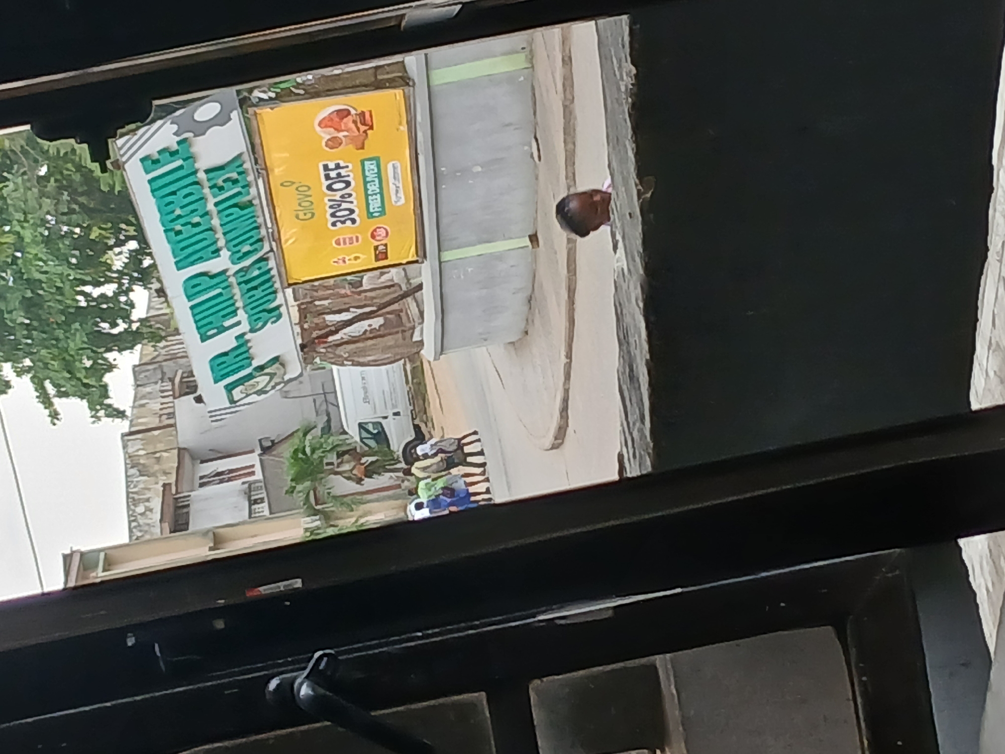 The image, seemingly taken from inside a building looking outwards through a window, shows a street scene. A sign for "DR. MUIR ADEBILE SPORTS COMPLEX" is prominently displayed in the scene, next to a yellow Glovo advertisement offering "30% OFF" with free delivery. Several children in casual clothing are walking on the sidewalk beside the Glovo advertisement. A light-colored van is parked nearby. A man's head is visible near the edge of the sidewalk. Buildings are visible in the background, amidst green trees. The setting is the city of Unknown, Unknown.