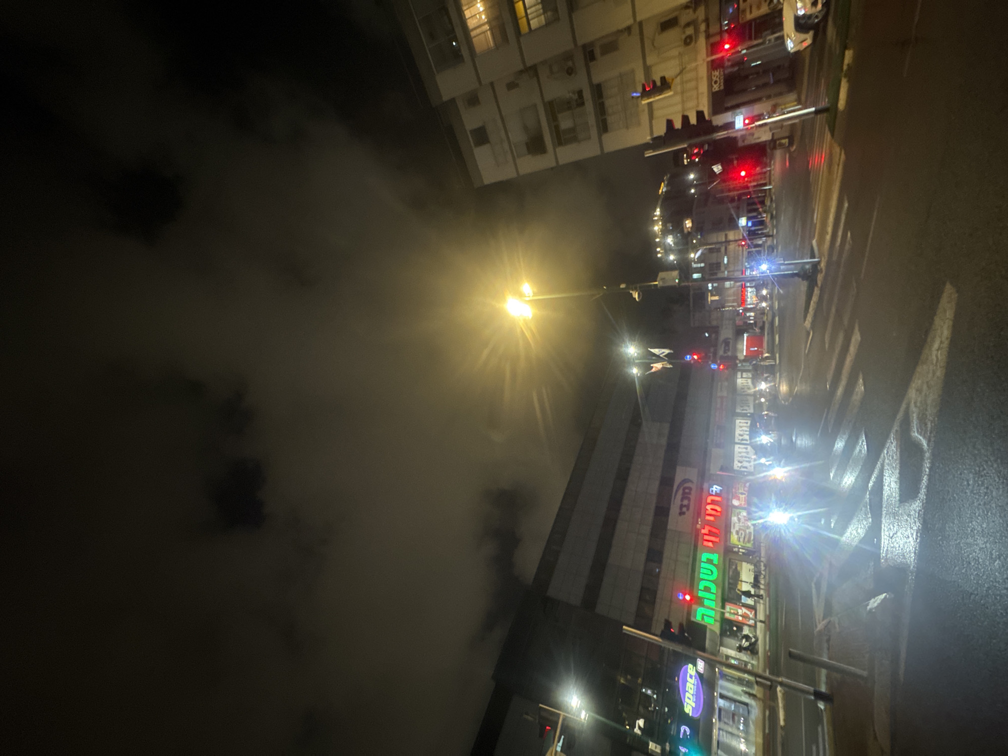 The image depicts a nighttime street scene in Ramat Gan, Israel. It shows a street with a crosswalk and what appears to be wet pavement. There is a building with the Hebrew sign "רמי לוי בשכונה" (Rami Levy BaShchuna) illuminated in green and red letters. Traffic lights and streetlights are visible, casting a glare. In the background, there are buildings with lit windows. The sky is dark, with possible cloud cover.