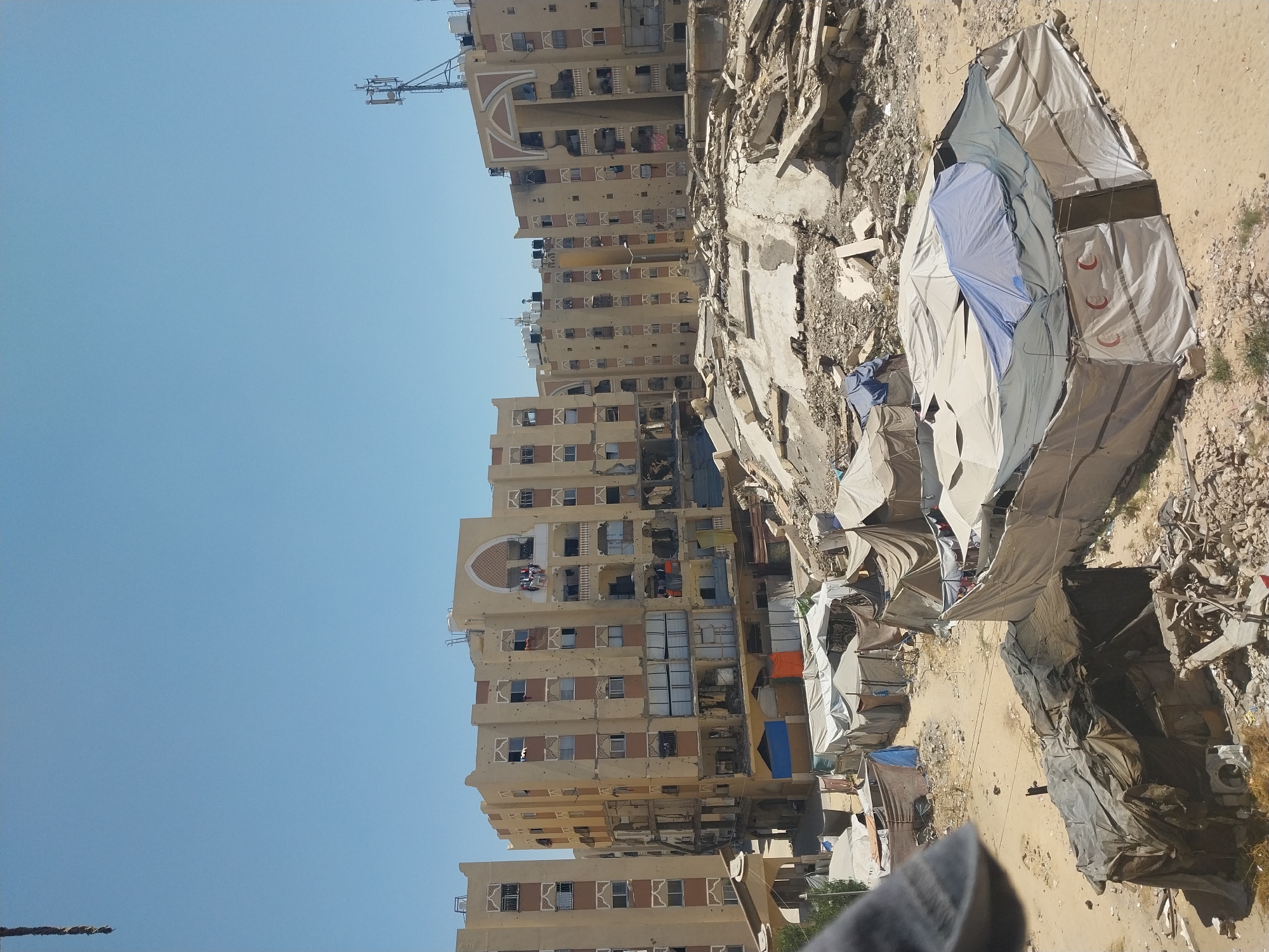 The image depicts a scene in Khan Yunis, Palestinian Territories, showing damaged multi-story apartment buildings adjacent to a collection of tents, indicative of a displacement camp. The buildings exhibit significant structural damage, with visible debris and holes in their facades, suggesting destruction. Water tanks are visible on the rooftops of the buildings. In front of the damaged buildings, a settlement of various tents has been erected. Some tents are made of white fabric, while others are constructed from darker materials. One prominent tent features the red crescent symbol, commonly associated with medical services or aid organizations. The ground appears to be sandy and uneven, scattered with rubble and debris from the damaged structures. The sky is clear and blue, suggesting a daytime setting. The overall environment conveys a sense of devastation and the presence of a temporary shelter community.