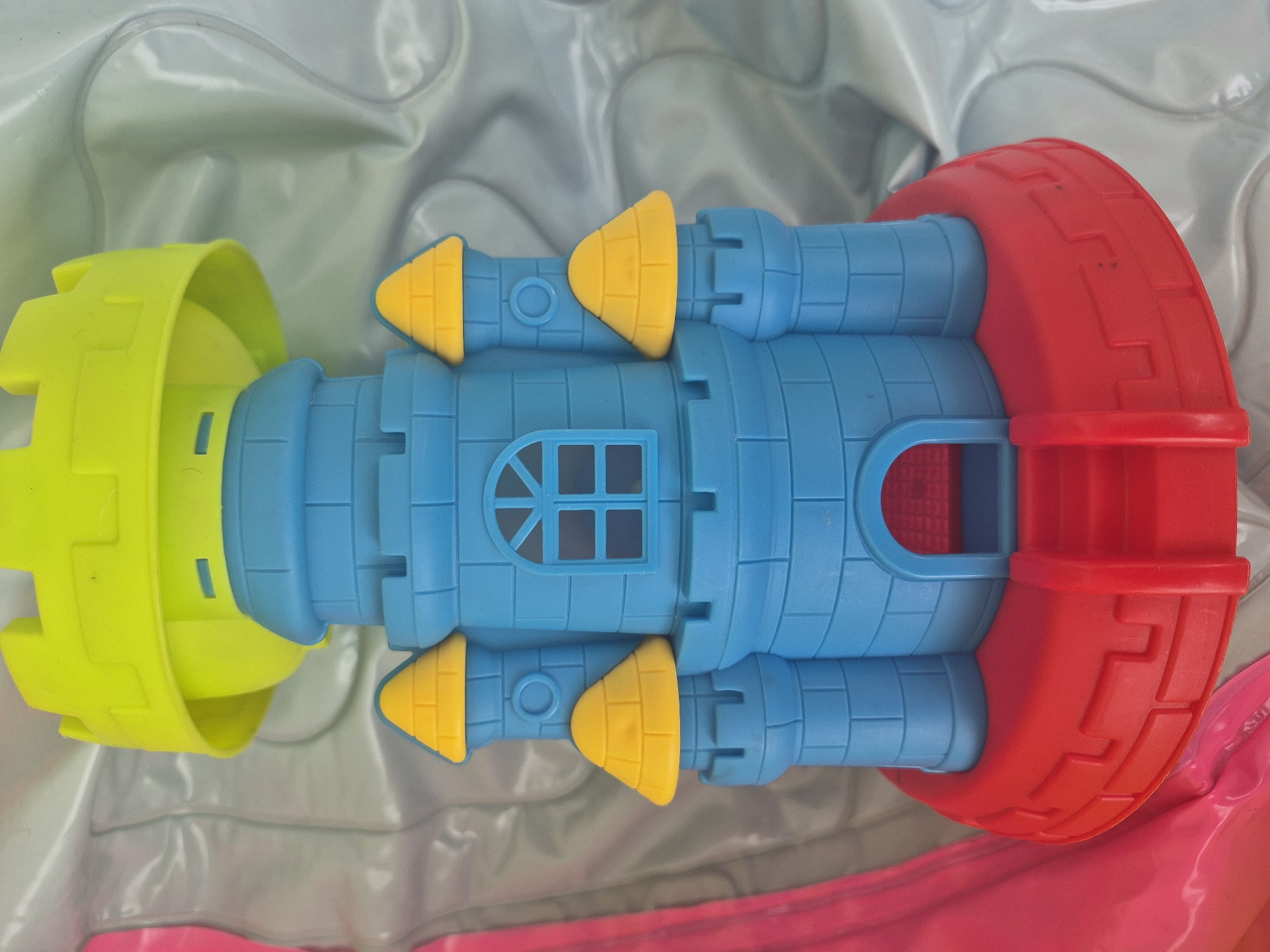 This media file features a vibrant, multi-colored plastic toy, likely a sandcastle mold or a children's play structure, placed on a soft, crinkled background. The toy is composed of several distinct sections.
The central part is a blue tower-like structure, molded with a detailed brick or block pattern. It includes several windows, one featuring a cross-hatched design and another with a simple arched opening. Smaller blue cylindrical sections extend from the main tower, each topped with a bright yellow conical spire.
On the left side, a large, lime green piece, shaped like a bucket with castellated edges, connects to the blue structure. To the right, a vivid red, semi-circular component, also textured with patterns, completes the toy. The bright colors—blue, yellow, lime green, and red—are characteristic of children's toys.
The toy rests on a light-colored, somewhat reflective plastic material, which appears to be crinkled, suggesting an inflatable surface like a pool liner or a play mat. A subtle hint of pink or magenta is visible along the bottom edge of the frame.
No activity or event is taking place; the image is a still life of the toy. The lighting is soft and diffused, providing no specific indications of time of day or weather. There is no visible text within the image. While the location is noted as Yerevan, Armenia, there are no visual cues in the photograph to confirm this context.