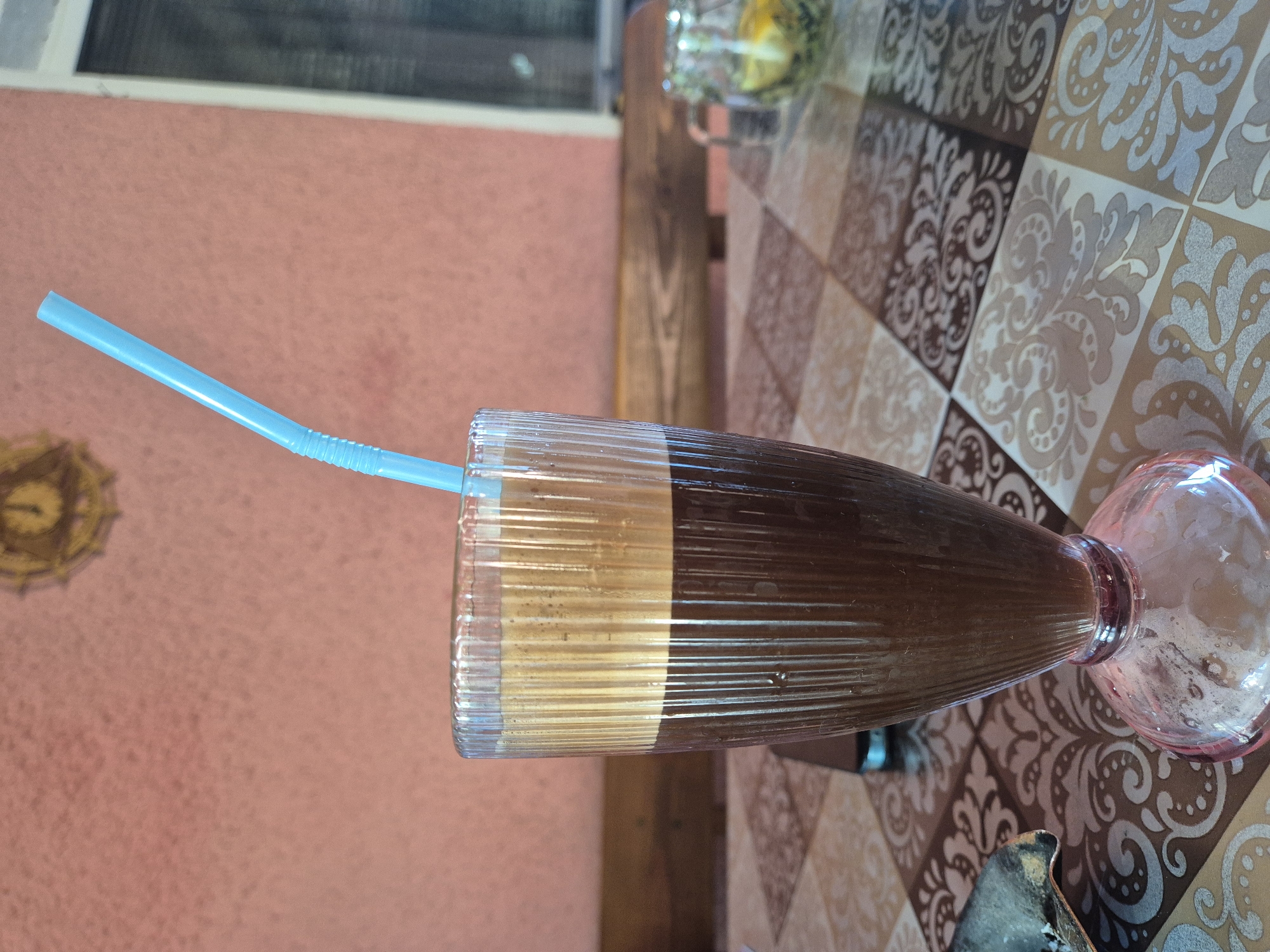 In an outdoor setting, a tall, fluted glass filled with a dark beverage topped with a thick layer of foam sits on a table. A blue plastic straw extends from the drink. The table is covered with a patterned tablecloth featuring squares with ornate designs in shades of brown and cream. In the background, a pink wall is visible, and to the right, a window with white trim and what appears to be shutters or blinds. A wooden bench is positioned behind the table. A small glass jar containing what looks like dried herbs and slices of lemon or lime is also present on the table. To the left, a small portion of a dark, metallic object is visible.