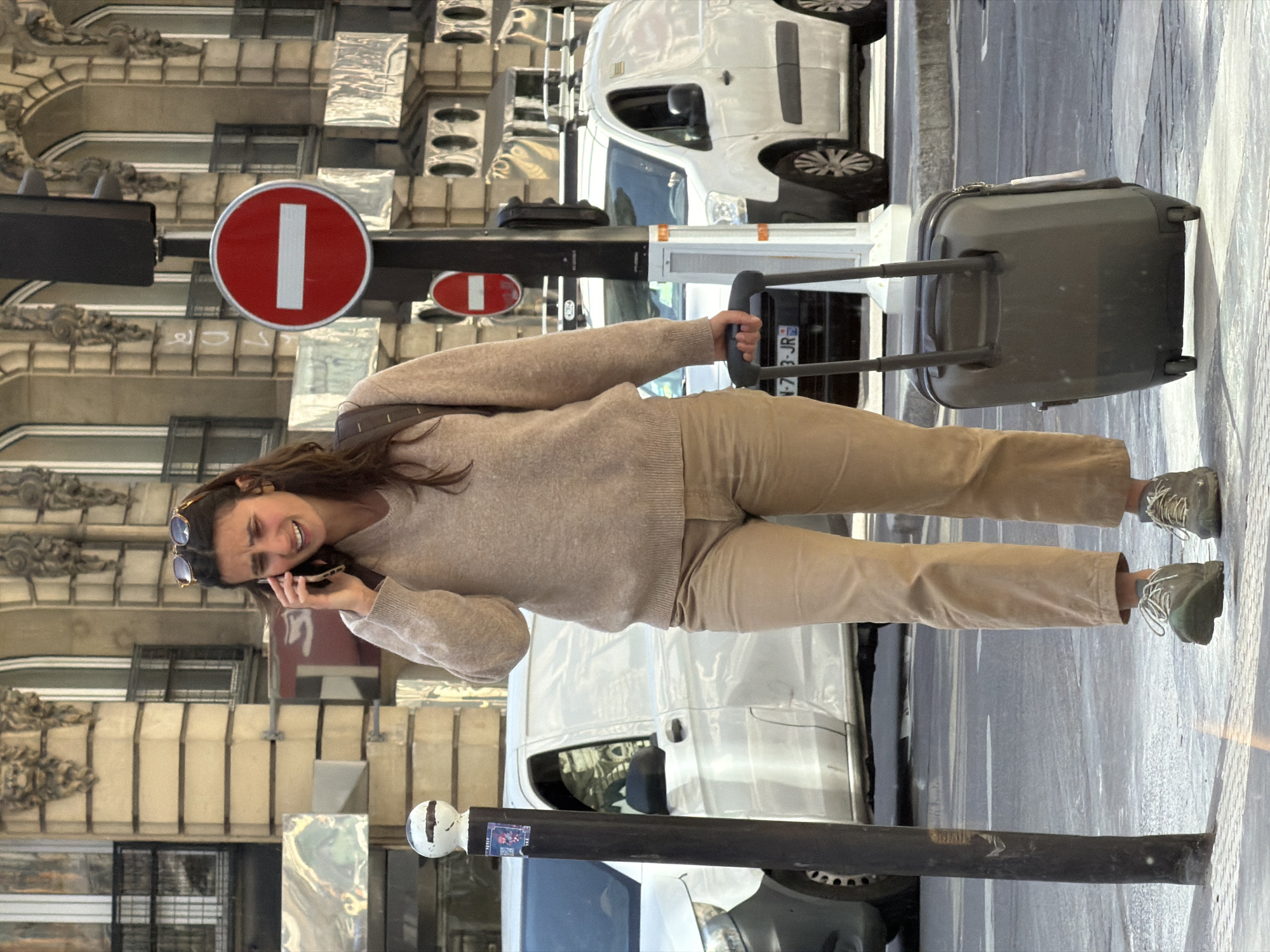 A woman, with sunglasses perched on her head, is walking on a street, pulling a rolling suitcase, and talking on a cell phone. She is dressed in light brown pants, a light brown sweater, and greenish-brown athletic shoes. She walks on the street near the sidewalk on a street in Paris. The street is narrow and lined with buildings of Parisian architecture. Traffic signs are visible, including a "Do Not Enter" sign. A white service van is parked nearby. The license plate of the van is visible. A lamp post is to her left. The street appears to be in a commercial or residential area of Paris.