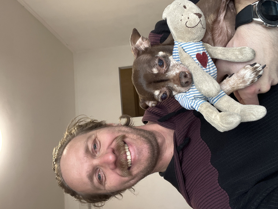 The scene depicts an adult male holding a small dog indoors, in what is stated to be Saint Petersburg, Russia.
The man, positioned on the left side of the frame, has light brown hair, a mustache, and a short beard. He is smiling broadly, revealing his teeth, and looking directly at the camera. He is wearing a long-sleeved hooded garment, which is dark burgundy with a ribbed texture on the upper section and transitions to black on the lower part, also with a ribbed texture. A black drawstring is visible near the hood. On his left wrist, a dark-colored smartwatch with a circular metallic bezel is worn.
He is holding a small, short-haired brown dog, consistent with a Chihuahua breed, with his left hand/arm supporting its body. The dog's face shows signs of age, with lighter fur around the muzzle and visible cloudiness in its eyes. The dog is holding a small, light-colored stuffed toy in its mouth. The toy appears to be a stylized animal, possibly a bear or rabbit, wearing a blue and white striped shirt adorned with a red heart appliqué on its chest. The toy has simple, embroidered facial features.
The background consists of a plain, light-colored wall, likely off-white or cream. A faint ceiling line and cornice are visible in the upper right. A dark brown rectangular opening, possibly a doorway, is partially visible in the mid-background to the left of the dog. The lighting appears to be artificial, originating from an overhead source not visible in the frame.