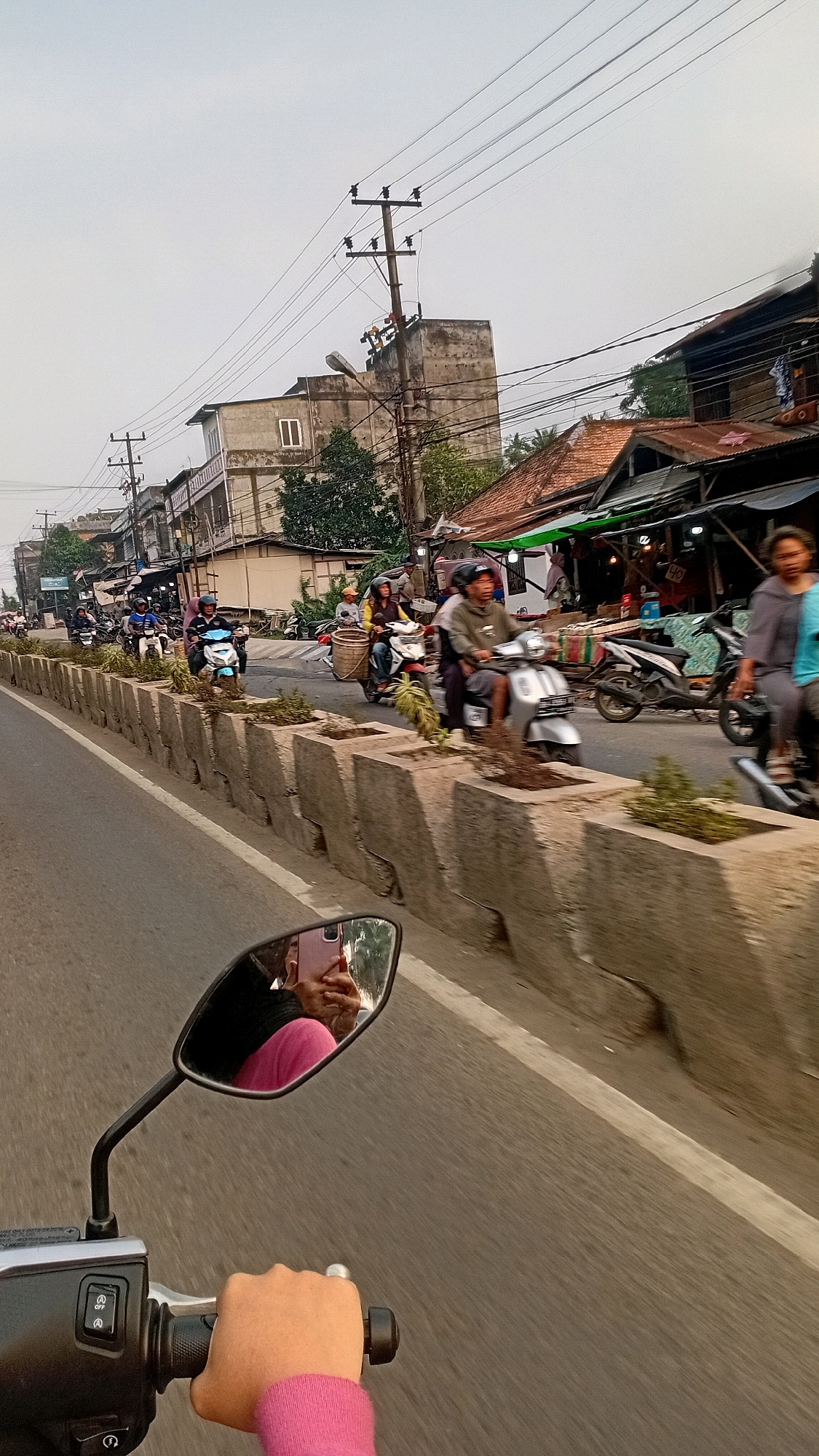 The image depicts a street scene in Jambi, Indonesia, with multiple motorcycles in motion. The perspective is from a motorcycle, showing the rider's hand on the handlebars and a rearview mirror reflecting the rider and their phone. The road is lined with concrete planters containing small plants, separating it from the sidewalk and buildings. On the sidewalk, there are several shops with awnings, and people are visible walking and riding motorcycles. Overhead, power lines and utility poles are prominent. The buildings appear to be a mix of older structures and newer constructions. The sky is overcast.