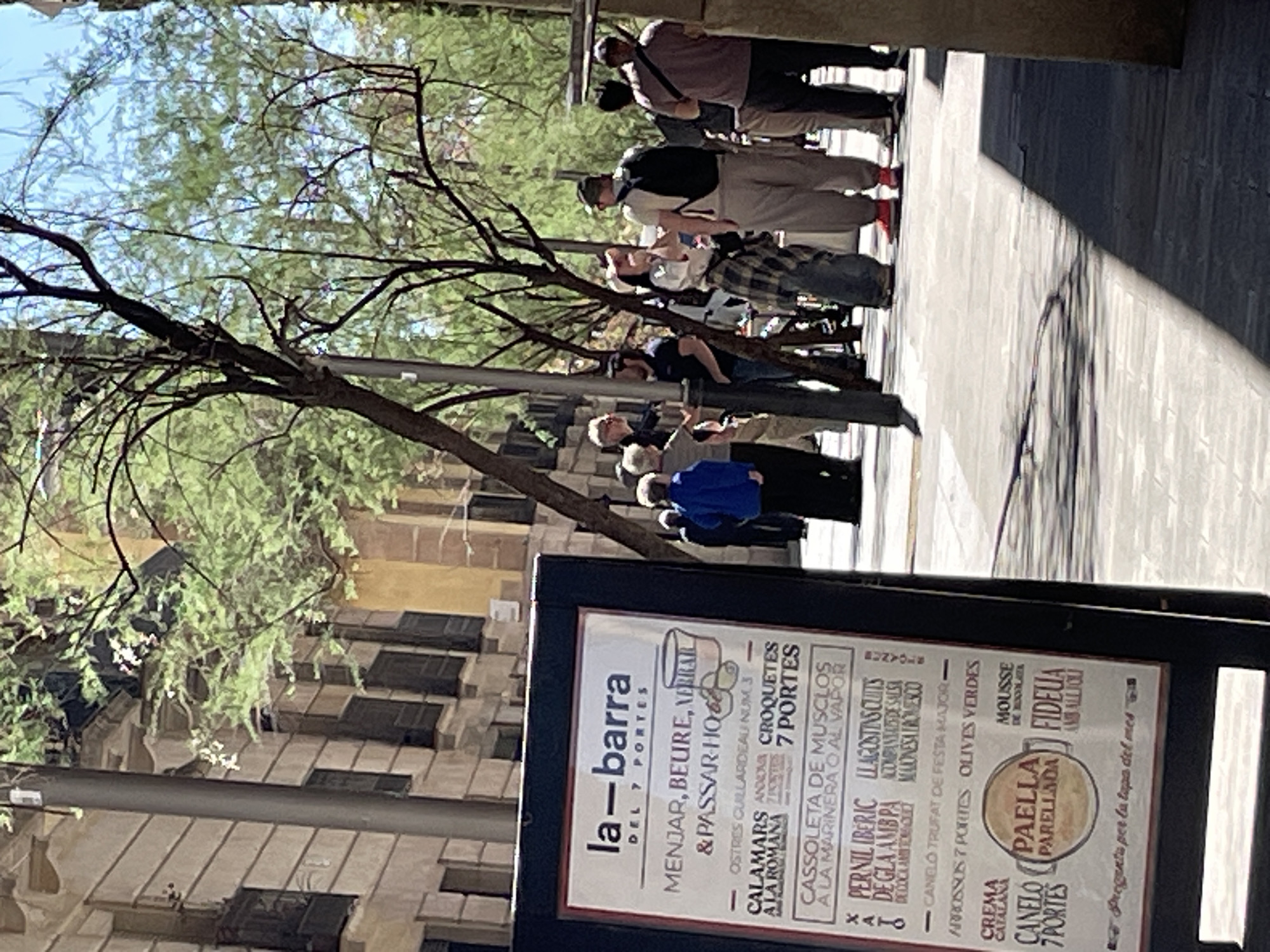 The image captures a vibrant daytime street scene in what appears to be Barcelona, Spain. A group of approximately ten people, dressed casually, are gathered or walking on a paved surface, some looking towards the background. They are positioned beneath the dappled shade of a tree with green foliage and prominent branches that extend across the upper left and center of the frame.
The setting features old, multi-story buildings with numerous windows in the background, characteristic of European urban architecture. The bright sunlight indicates daytime, casting strong shadows on the ground and creating a well-lit environment.
In the foreground, a black sandwich board sign for a restaurant named "la-barra DEL 7 PORTES" is prominently displayed. The sign, written in Catalan/Spanish, lists various traditional dishes and offerings. Visible text includes "MENJAR, BEURE, & PASSAR-HO" (Eat, Drink, & Have a good time), "OSTRES CUALLARDEAU NUM 3", "CALAMARS A LA ROMANA", "CROQUETES 7 PORTES", "CASSOLETA DE MUSCLOS A LA MARINERA O AL VAPOR" (Mussel casserole), "PERNIL IBERIC DEGLA AMB PA" (Iberian ham with bread), "CANELO TRUFAT DE FESTA MAJOR" (Truffle cannelloni), "ARROSSOS 7 PORTES", "OLIVES VERDES", "CREMA CATALANA", "MOUSSE DE XOCOLATA", "PAELLA PARELLADA", and "FIDEUA AMB ALLIOLI" (Noodle paella with aioli). It also mentions "Pregunta per la tapa del mes" (Ask for the tapa of the month). The overall impression is a bustling, sunny day outside a traditional eatery.
