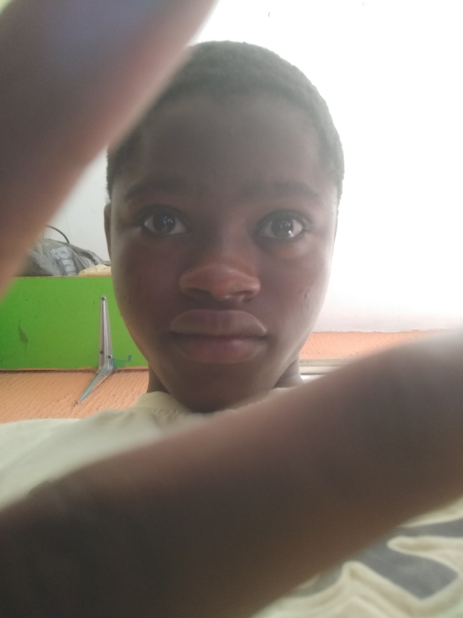 A close-up selfie of a young Black person with short hair. The person is wearing a light-colored shirt. Part of the person's arm is visible in the foreground. A green object and a silver object with two pointed ends are visible in the background. The location is indoors, presumably in Shangisha, Nigeria.