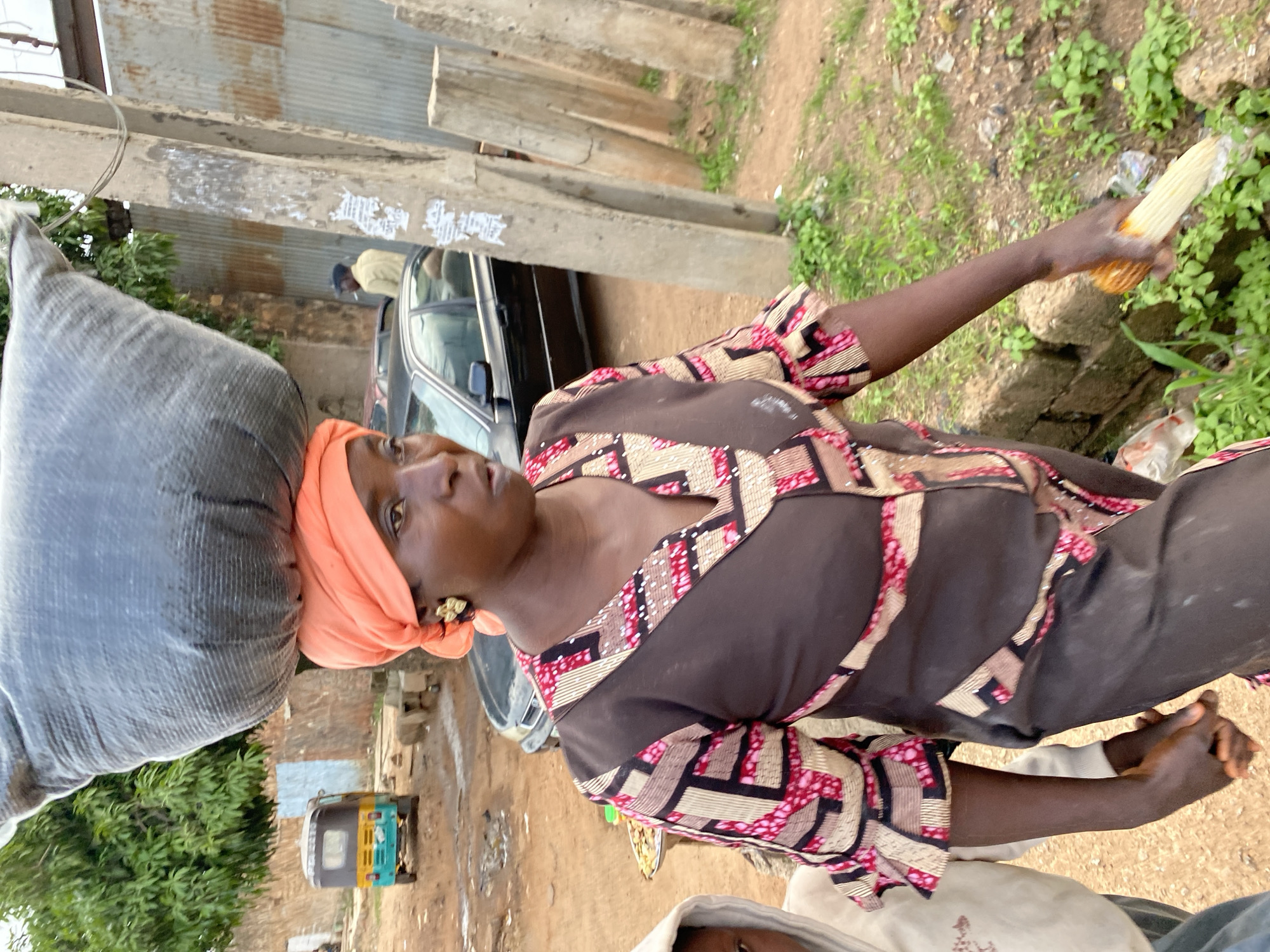 A woman, wearing a brown and pink patterned dress and an orange headscarf, carries a large sack on her head. Her left arm is bent at the elbow, and she holds a cob of corn in her hand. She is standing outdoors in what appears to be a market or roadside setting. In the background, a dark-colored car is parked, and a small, colorful auto-rickshaw is visible. The ground is dirt, and there are some green plants and debris scattered around. To the left, there is a corrugated metal structure and some concrete beams. The lighting suggests it is daytime. The woman's facial expression is neutral, and her gaze is directed slightly to the left. The overall scene depicts everyday life in a busy environment, likely in Bukuru, Nigeria.