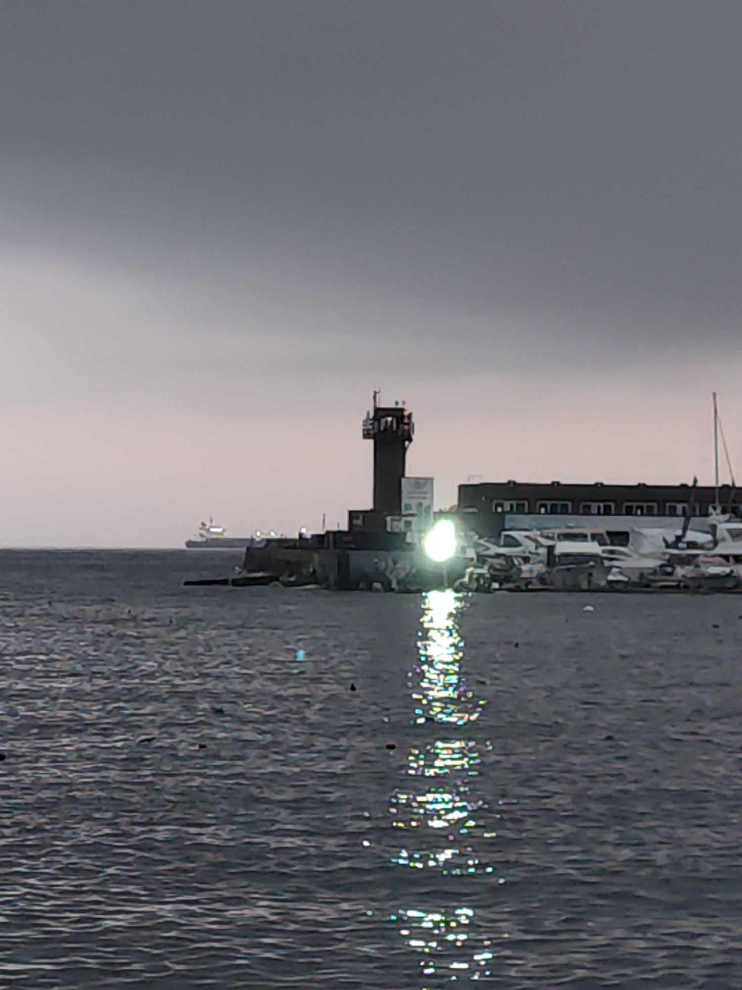 This is a daytime, slightly overcast, long-shot photograph of a harbor area in Vladivostok, Russia. A lighthouse, a building, and several boats are visible. In the foreground, dark, rippling water reflects the sky and a bright light source, possibly from the lighthouse, creating streaks of shimmering light on the surface. In the mid-ground, a dark, slender lighthouse stands on a pier. Beside it is a long, dark building with multiple windows. Several boats, including yachts and smaller vessels, are docked in front of the building. In the background, a large cargo ship is visible on the horizon. The sky is a uniform shade of gray, indicating an overcast or cloudy day.