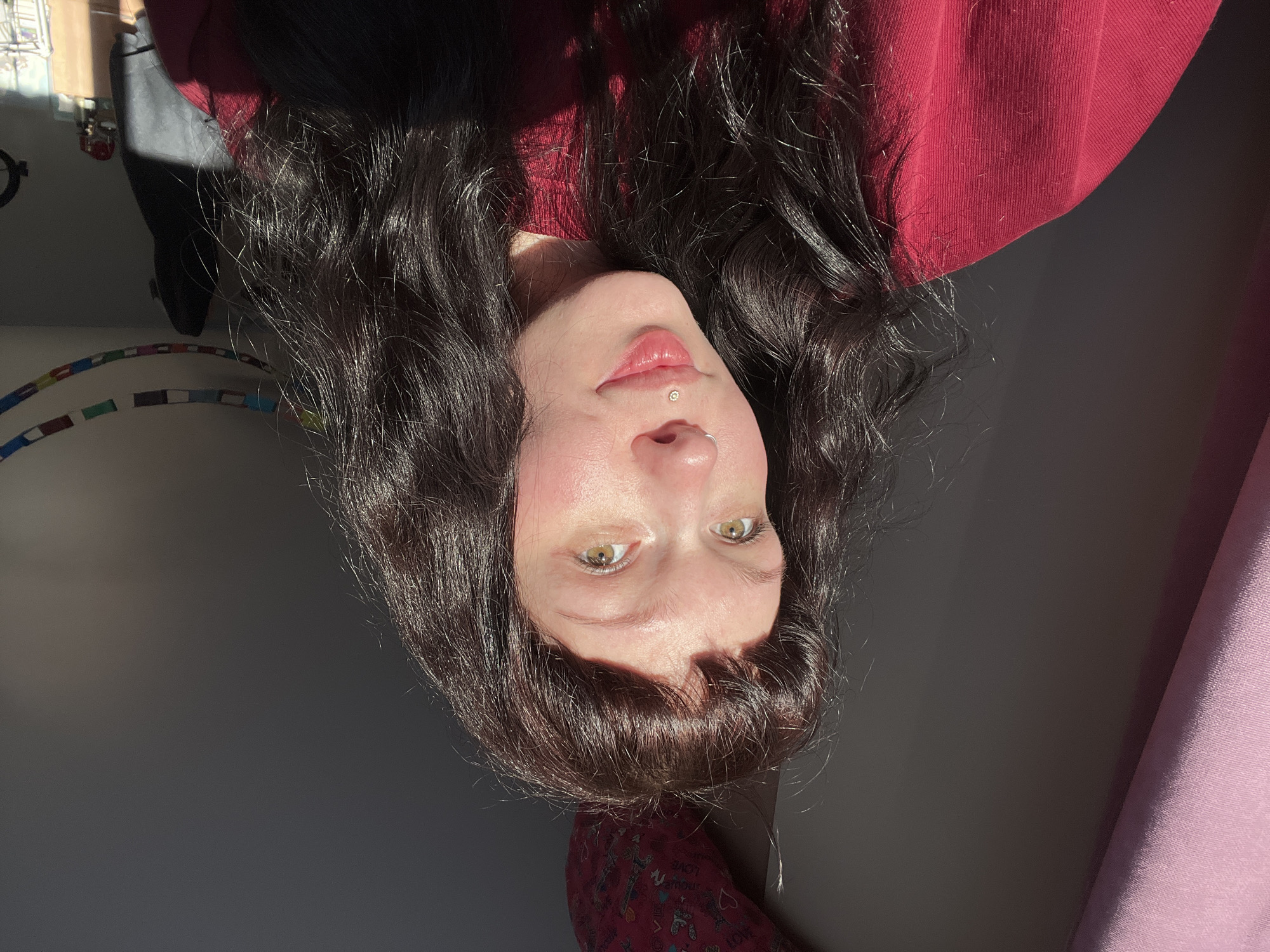 An image taken in Chaikovsky, Russia, features a young woman positioned centrally, her torso and head visible. She has dark brown, wavy hair with bangs across her forehead, and light skin with a flush on her cheeks. Her eyes are greenish-hazel. A silver hoop nose ring is present on her left nostril, and a gold-colored stud with a clear gem is in a philtrum piercing above her upper lip. She wears a red long-sleeved garment. Her face is illuminated from the right by direct sunlight, casting shadows on the left side of her face and the background. She looks slightly to the viewer's left with a subtle smile.
The background on the left features a portion of a pink fabric or curtain. Above her head, a dark red fabric object, possibly a pillow, displays patterns including what appears to be hearts, Eiffel Towers, and text like "LOVE." The wall behind her is a light gray color. In the upper right background, a multicolored string of decorative flags or bunting is visible. Further to the right, a dark object resembling a garment bag or instrument case hangs, and a circular object is mounted on the wall. Sunlight also illuminates a small section of furniture or shelves on the far right.