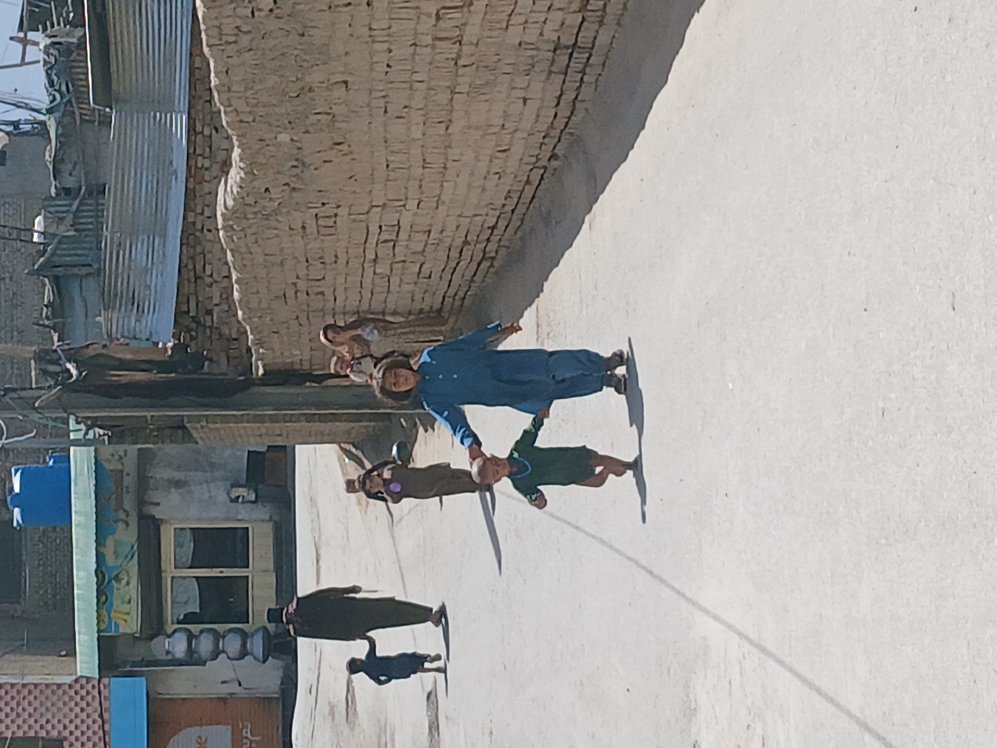 The image depicts a street scene in Quetta City Tehsil, Pakistan. A group of children are visible, with two being the primary subjects. One child, wearing a blue outfit, holds the hand of another, who is wearing green, and together they are doing an activity like a cartwheel. Other children stand near a brick wall. Along the street are buildings of brick or corrugated metal. Shadows suggest bright sunlight.