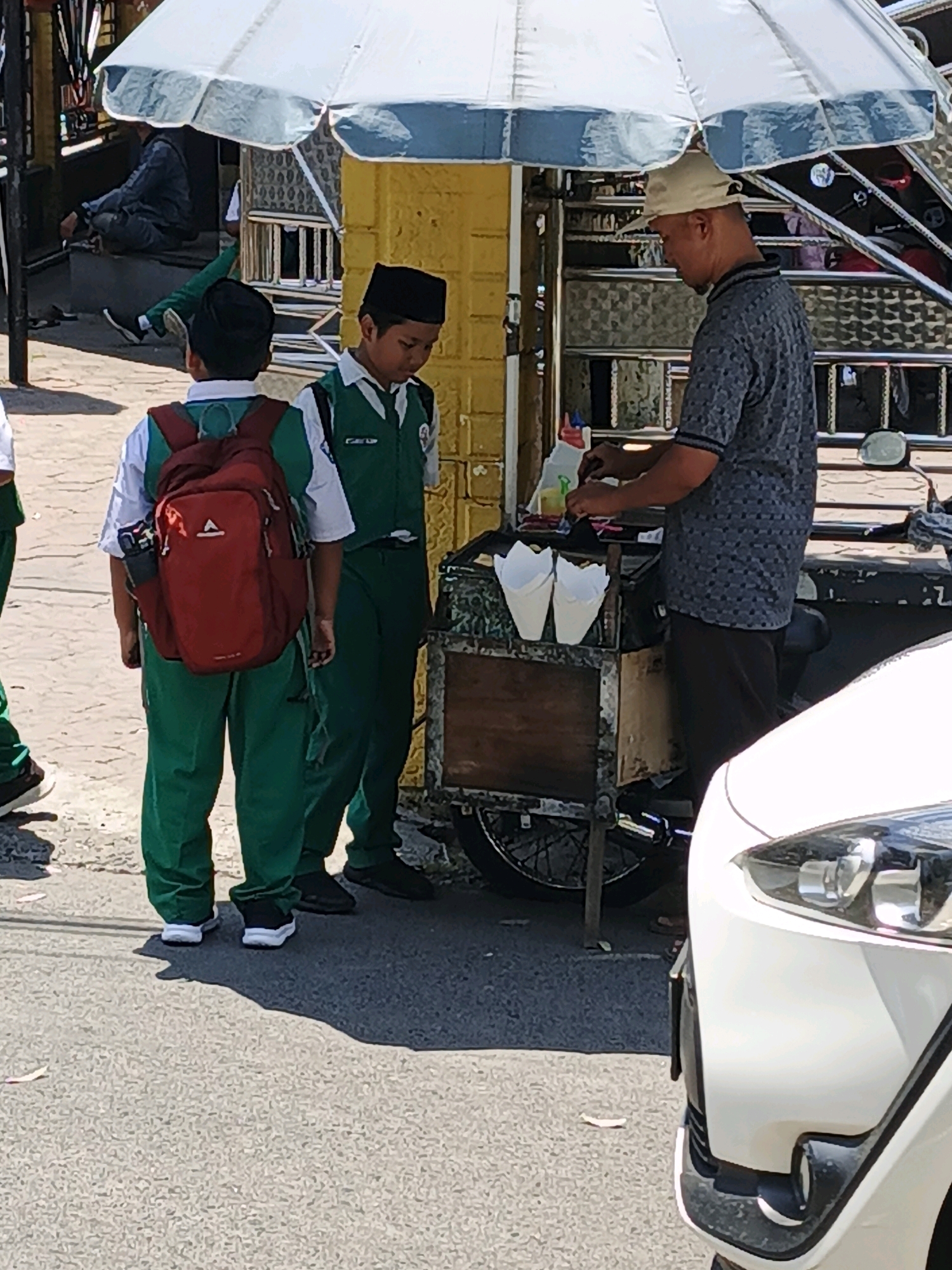 In the city of Ketegan, Indonesia, a street scene unfolds beneath a white umbrella. Two young boys, clad in school uniforms consisting of white shirts, green vests, and green pants, stand near a food cart. The boy on the left wears a red backpack. The boy on the right is looking down. A man wearing a dark patterned shirt and a light-colored cap operates the food cart, which is mounted on a wheeled base. He is in the process of serving customers. The cart contains white paper cones and what appears to be condiments. In the foreground, the front of a white car is visible. The environment suggests a sunny day, with shadows cast on the paved street.