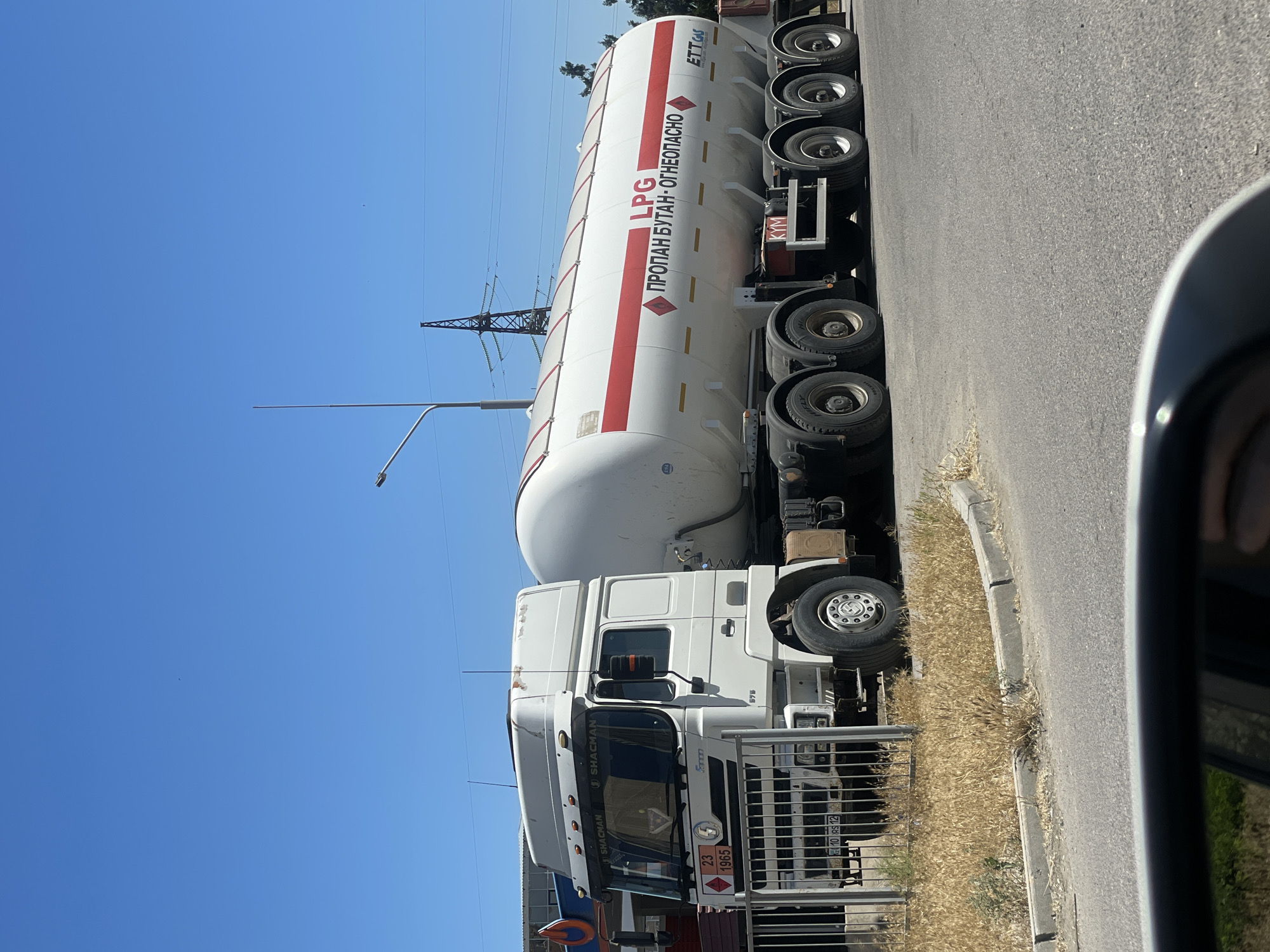 The image shows a white semi-trailer truck parked on the side of a road under a clear blue sky. The truck is transporting a large white cylindrical tank with red stripes, labeled "LPG" and "ПРОПАН БУТАН - ОГНЕОПАСНО" (Propane Butane - Flammable) in Russian. The truck itself has "SHACMAN" written on the cab. There are multiple wheels visible on the trailer and the truck. Electrical pylons and a street light are visible in the background, suggesting an urban or industrial setting. The scene appears to be taking place during the daytime, with bright sunlight. The overall impression is a utilitarian scene of a fuel transport vehicle at rest.