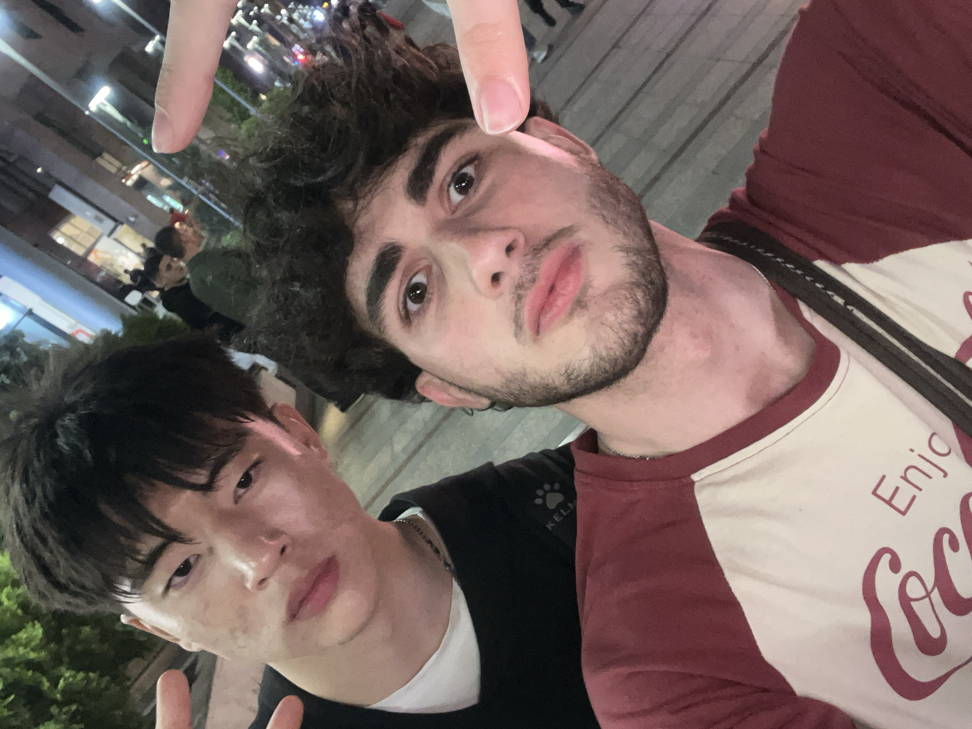 Two young men are posing for a selfie at night. The man on the right is fair-skinned with dark curly hair and a short beard. He's wearing a maroon and white shirt. The man on the left has dark hair and is wearing a black jacket. One hand is raised above the first man’s head and the other is making a peace sign. The background shows streetlights and other people, suggesting an urban setting. The city of Yerevan, Armenia is not apparent from the image and location cannot be determined.