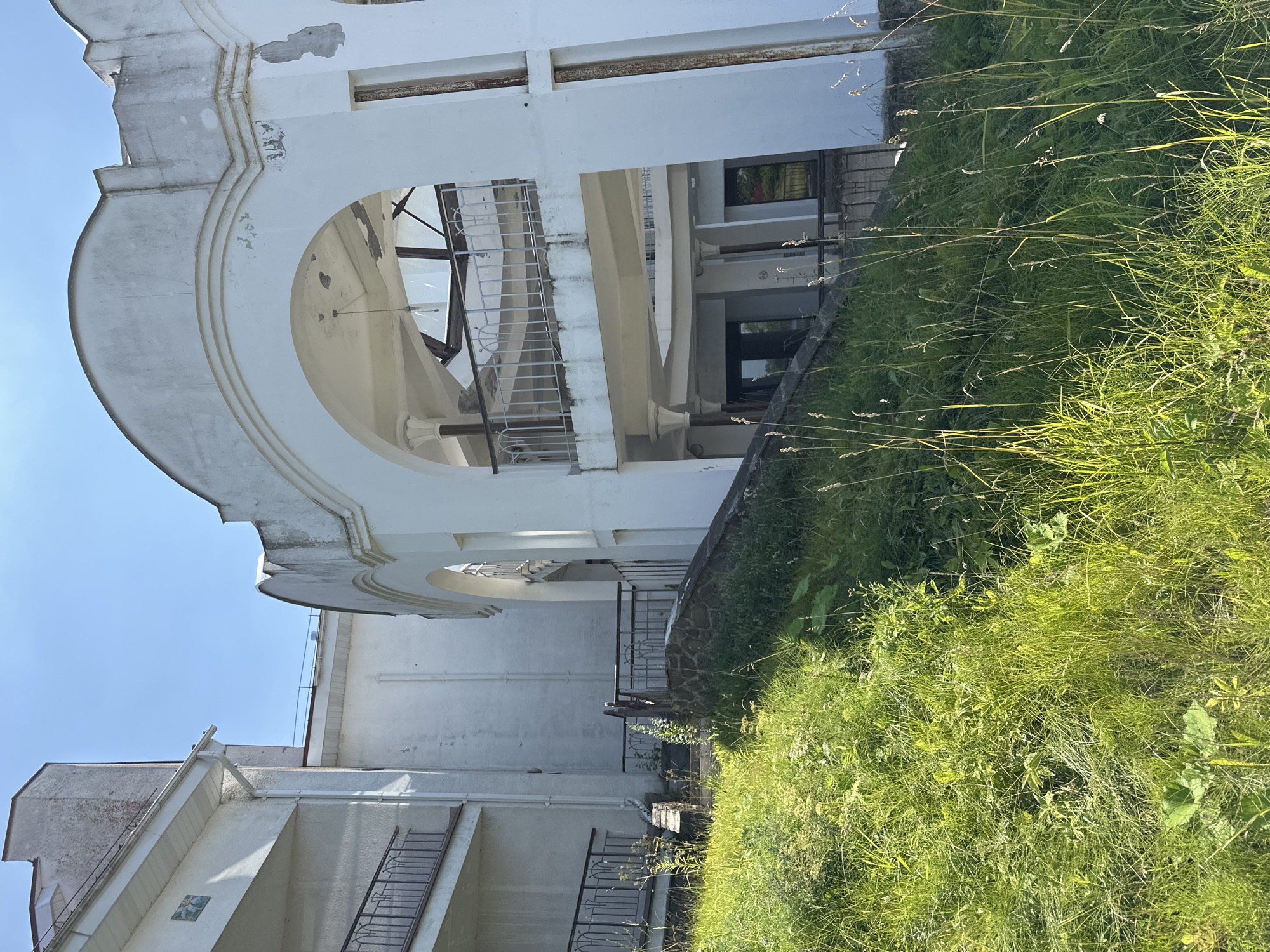 This is a low-angle, medium shot of an overgrown green slope leading up to an abandoned, white, multi-story building under a clear blue sky. The building's architecture features arches, balconies with ornate railings, and concrete columns. The facade shows signs of disrepair, with peeling paint and some rusting metal elements. The foreground is dominated by tall, green grass and weeds, obscuring a stone pathway and railing leading towards the building. The scene suggests an outdoor, possibly rural or park-like setting, given the abundant vegetation and the neglected state of the structure.